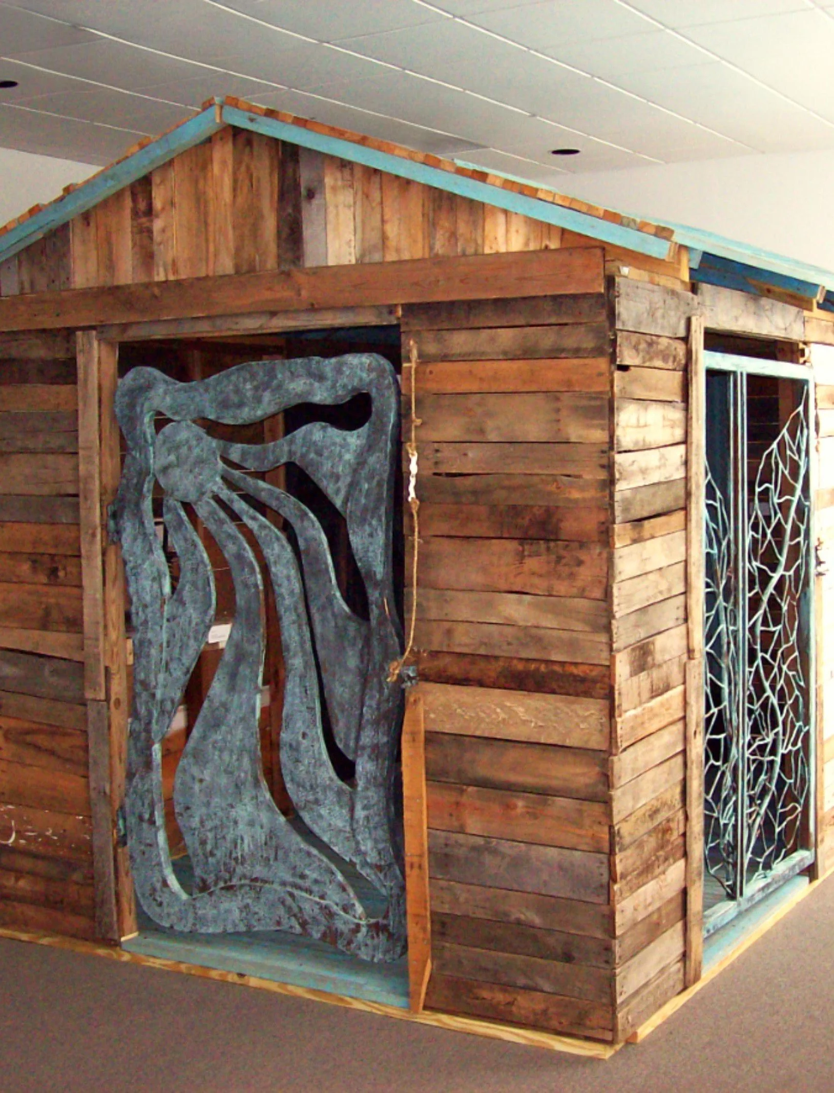 A wooden art installation or display with a cutout of a stylized whale, located indoors on a carpeted floor, with a white ceiling and decorative window panels nearby.