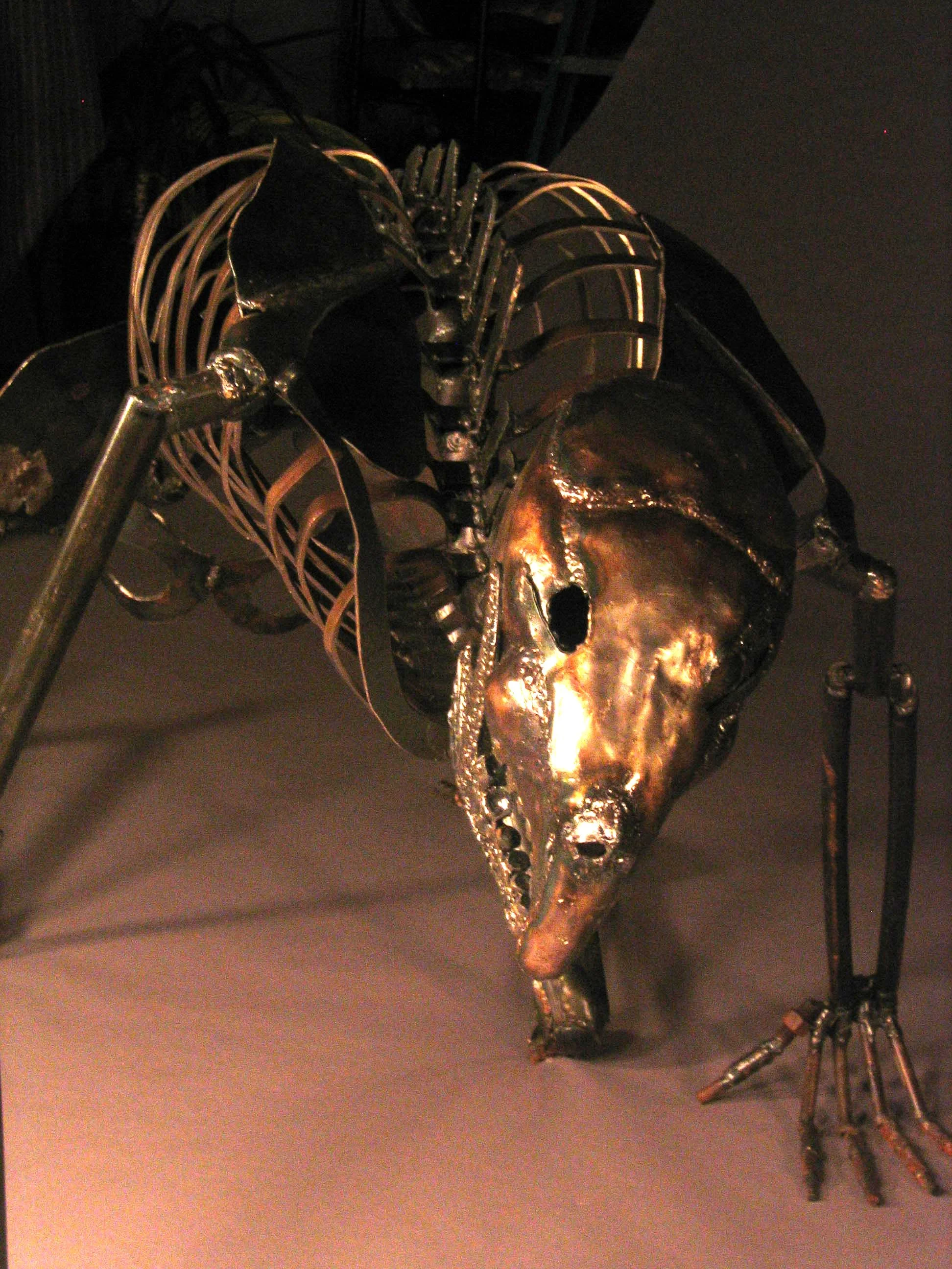 A metallic sculpture of an animal skull with horns and mechanical elements, displayed on a stand against a dark background.