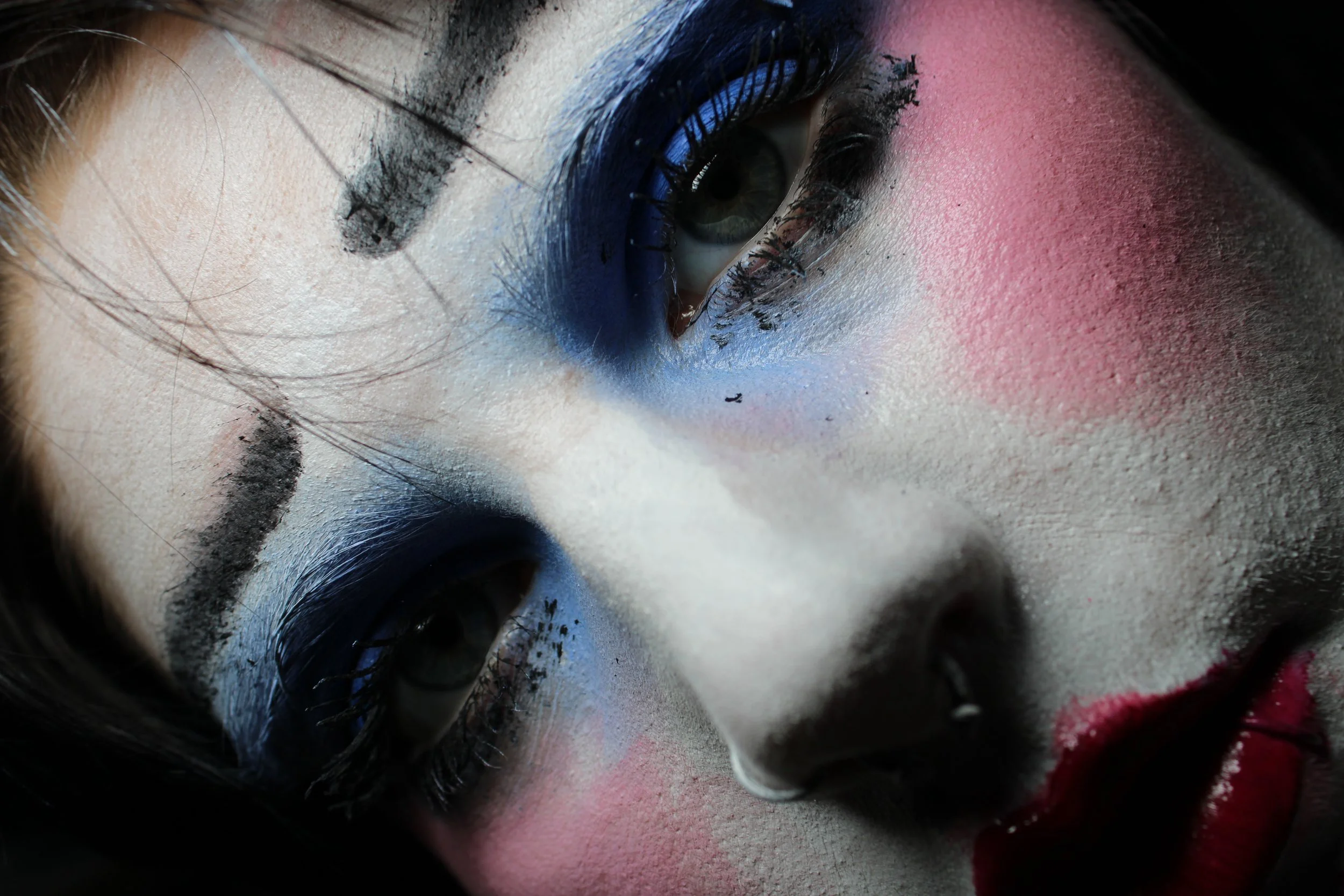Close-up of a person's face with smeared colorful, clown-like makeup and face paint, using pink, white, and black , and black eyeliner and mascara.