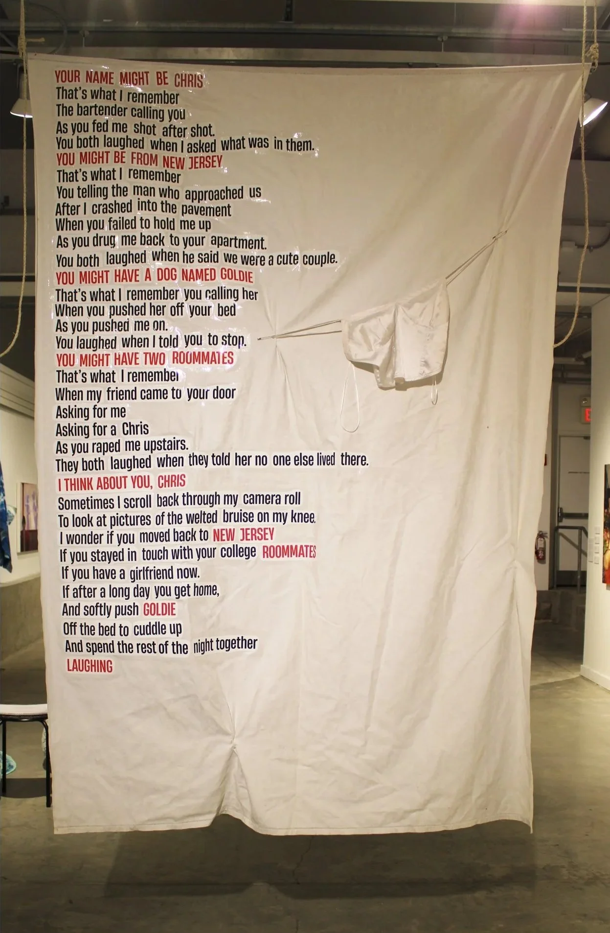 A large white fabric banner with printed text, hanging from the ceiling with cords on the sides. The banner contains poetic verses in black and red text, mentioning names like Chris, Goldie, and New Jersey, with emotional and reflective themes.