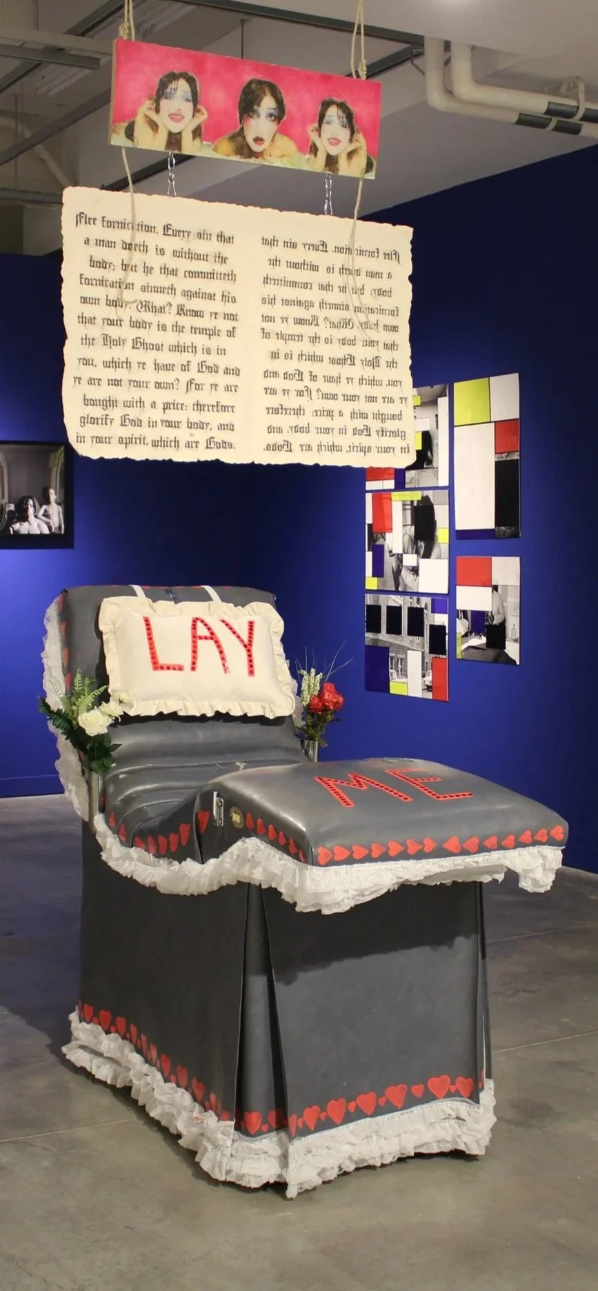 An art installation features a vintage hospital gurney decorated with a black cloth covered with white lace and red heart accents. The pillow on the gurney spells out "LAV" in red letters, and the foot of the gurney spells out "ME" in red letters. A 