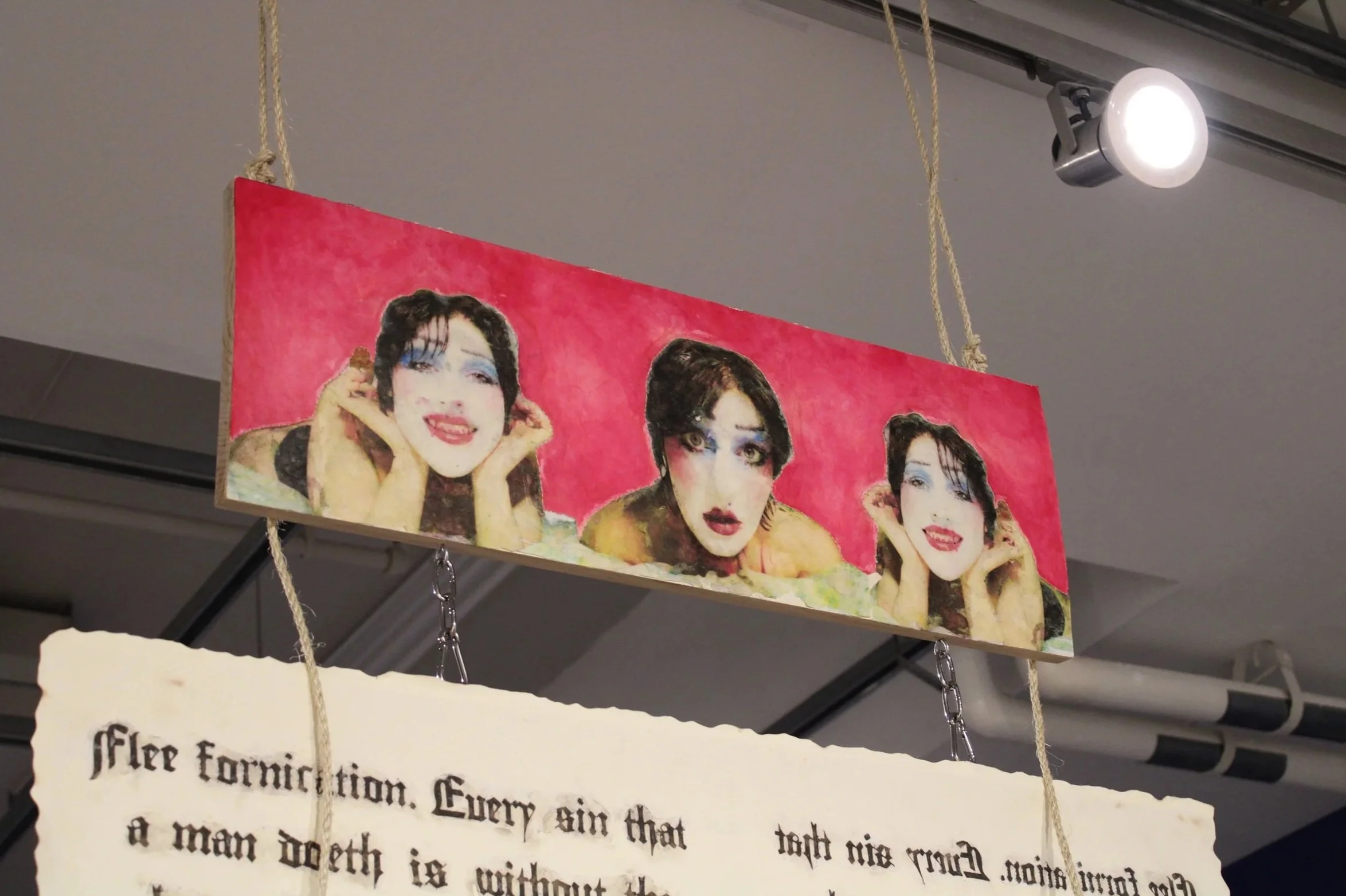 A colorful illustrated banner featuring multiple portraits of a woman with dark hair, red lipstick, and blue eye makeup, set against a pink background, hanging from the ceiling with ropes.