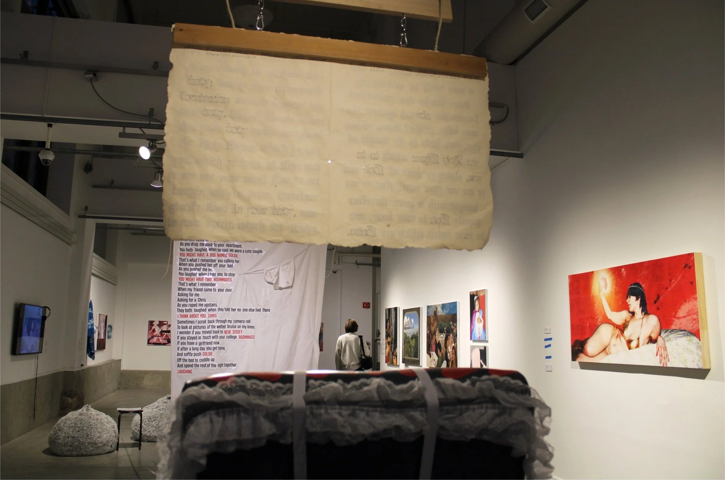 An art gallery with various paintings on the walls, a large hanging paper with text, and people observing the art. The gallery has industrial lighting and minimalist decor.