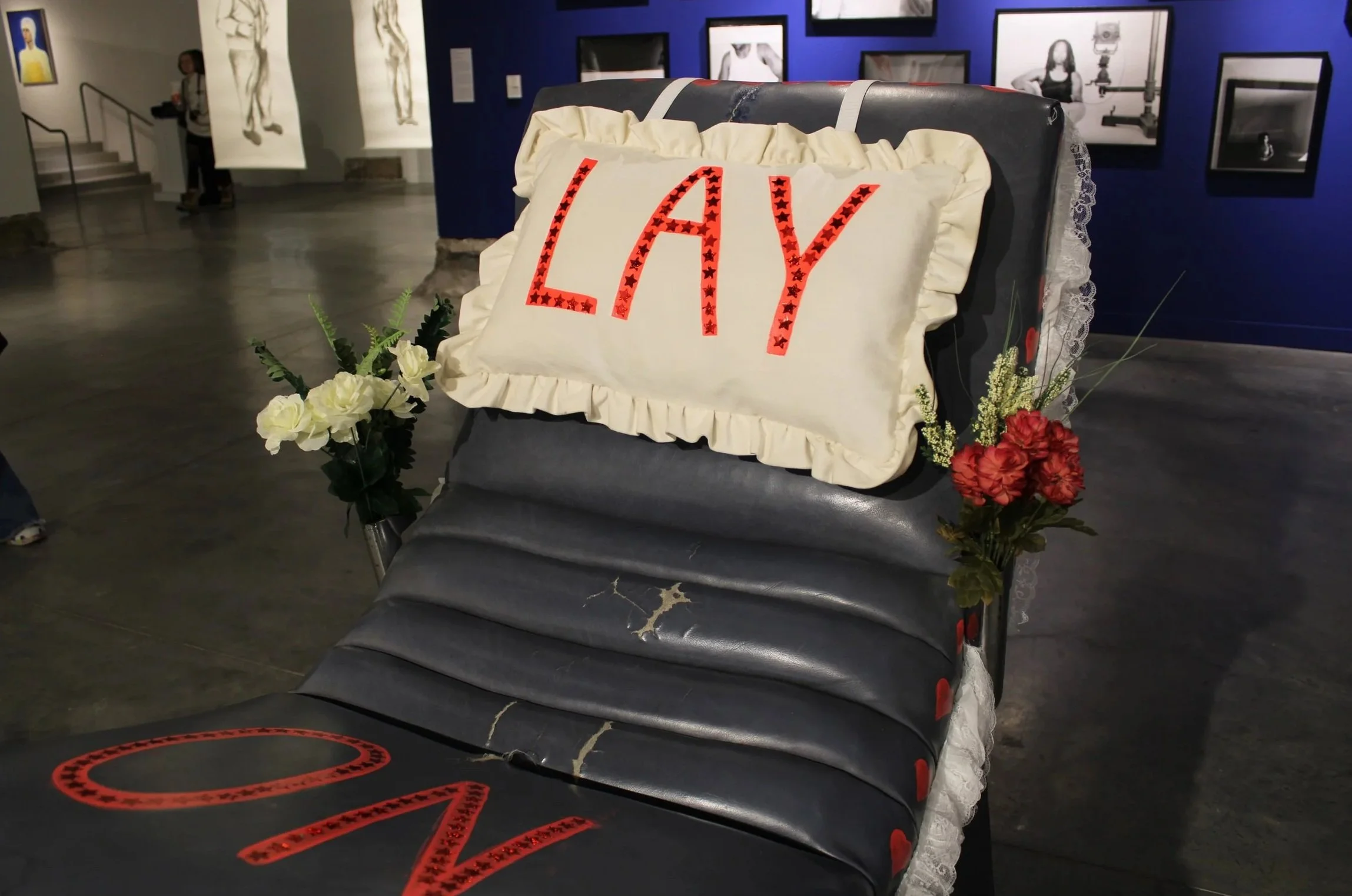 A large, decorated cake with the word 'LADY' written in red with black stars on top, placed on top of a black, worn-out, padded bench in an art gallery with framed black and white photos on the walls and floral arrangements on either side of the cake