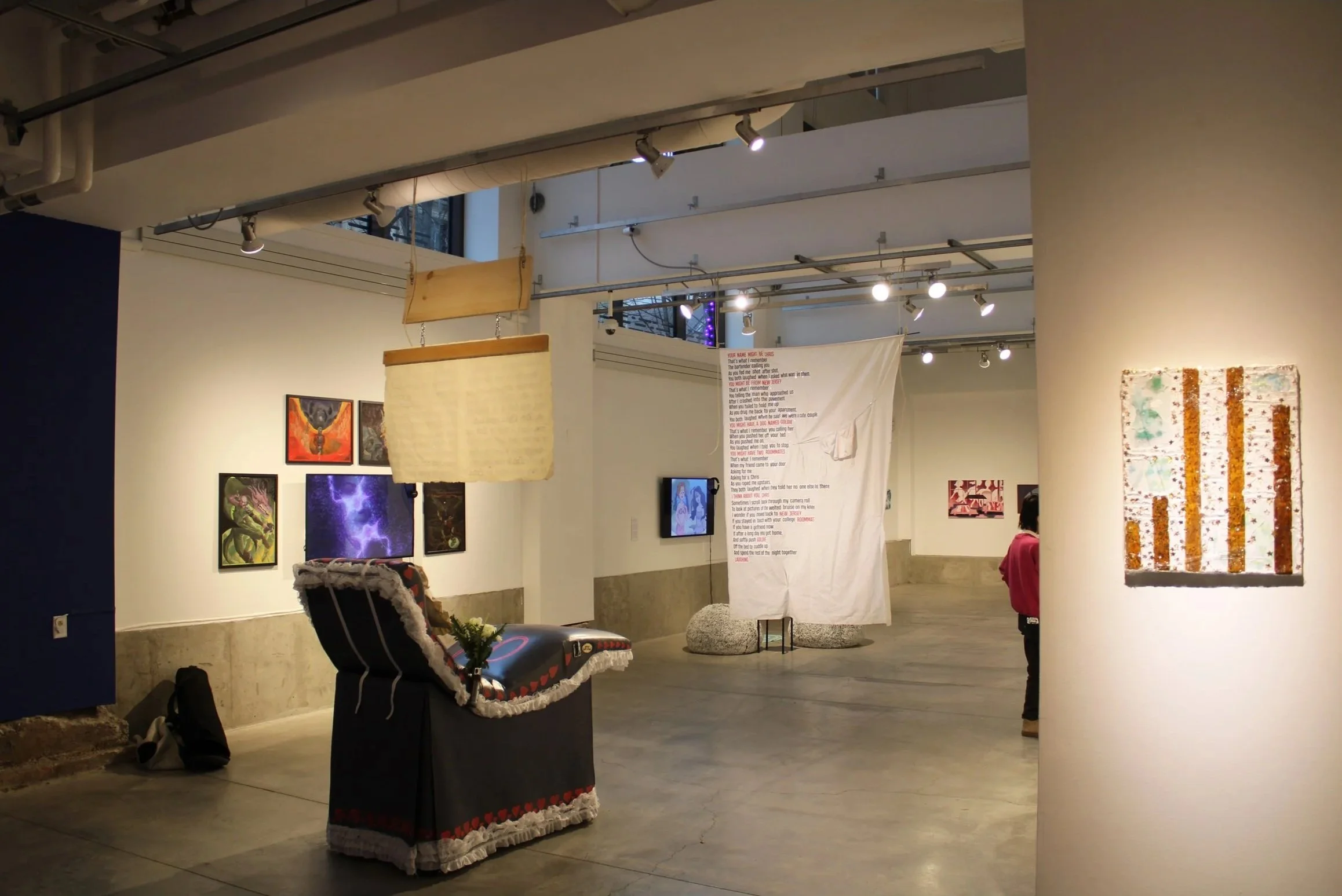An art gallery with various paintings hanging on the walls. A wooden hanging sign and fabric display with text are visible, and a person in a red jacket is partly seen in the background. The gallery has bright overhead lighting and a modern, industri