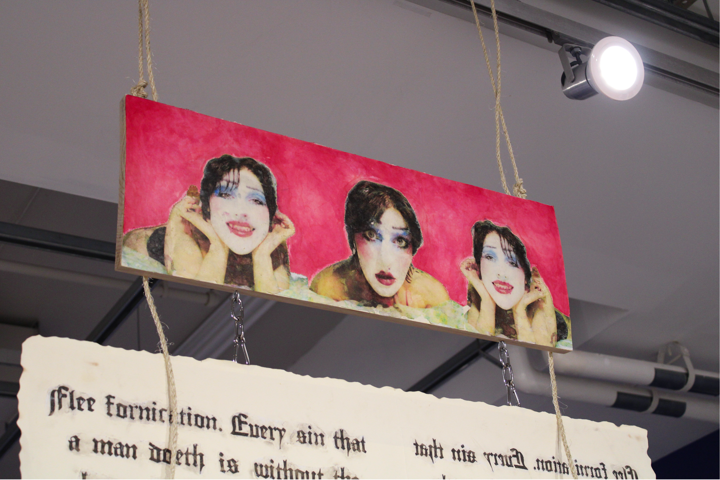 A hanging banner features repeated images of a person with dark, styled hair, makeup, and a playful expression, set against a pink background. Below the images, part of a white paper with black text is visible.