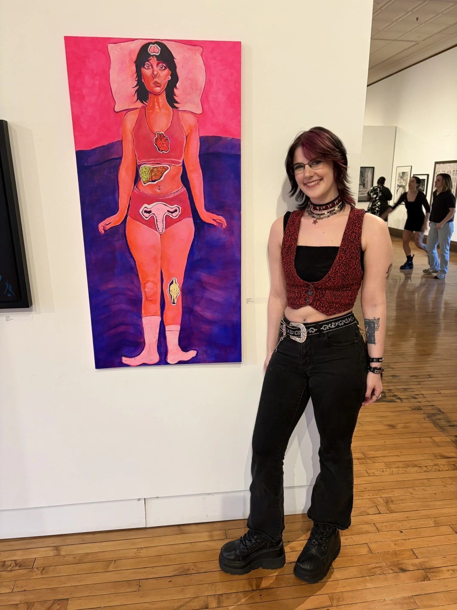 A person with dark, styled hair and glasses smiling at an art gallery, standing in front of a colorful painting of a person lying on a bed with visible internal organs.