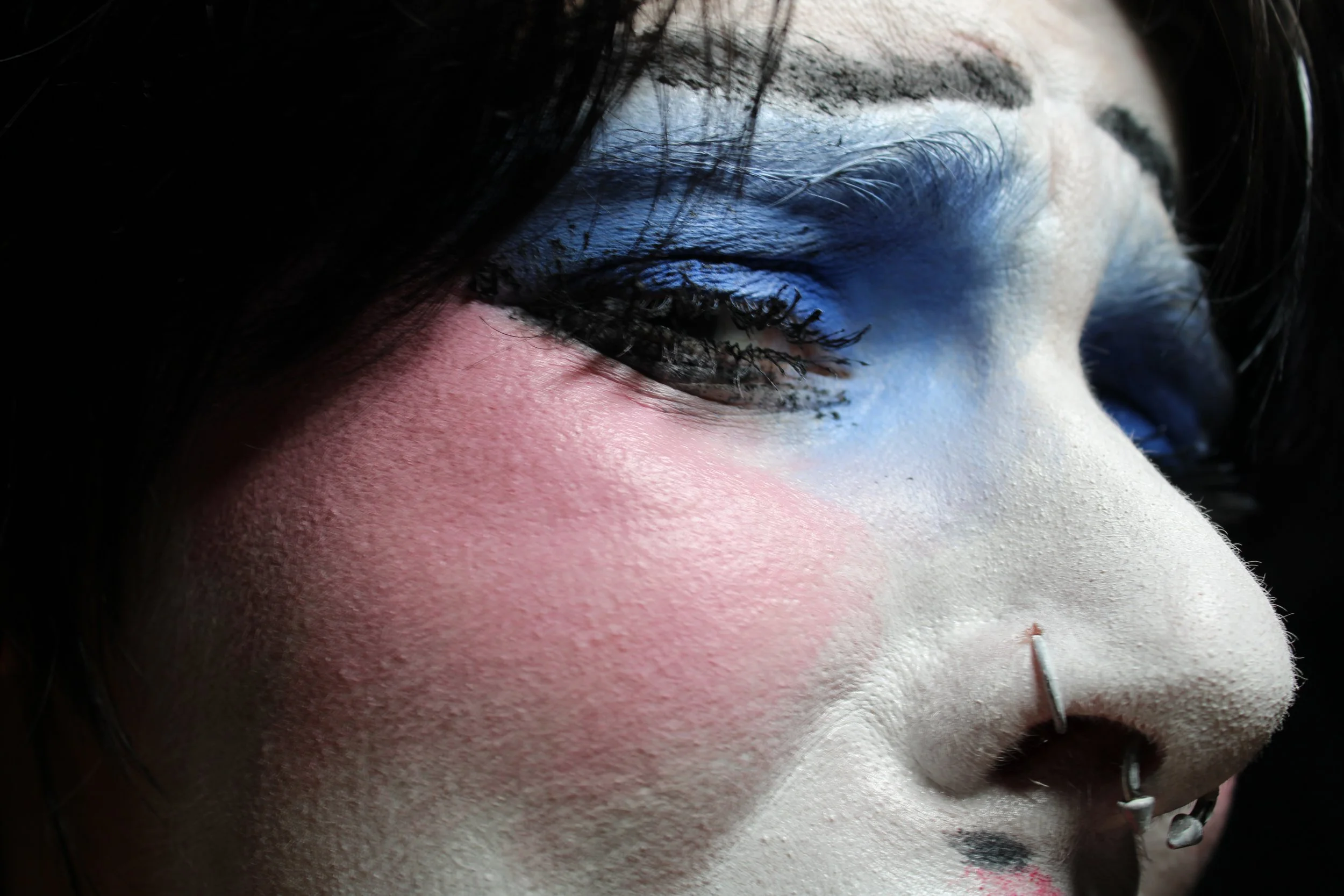Close-up of a person's face with artistic makeup, dark hair, and facial piercings.
