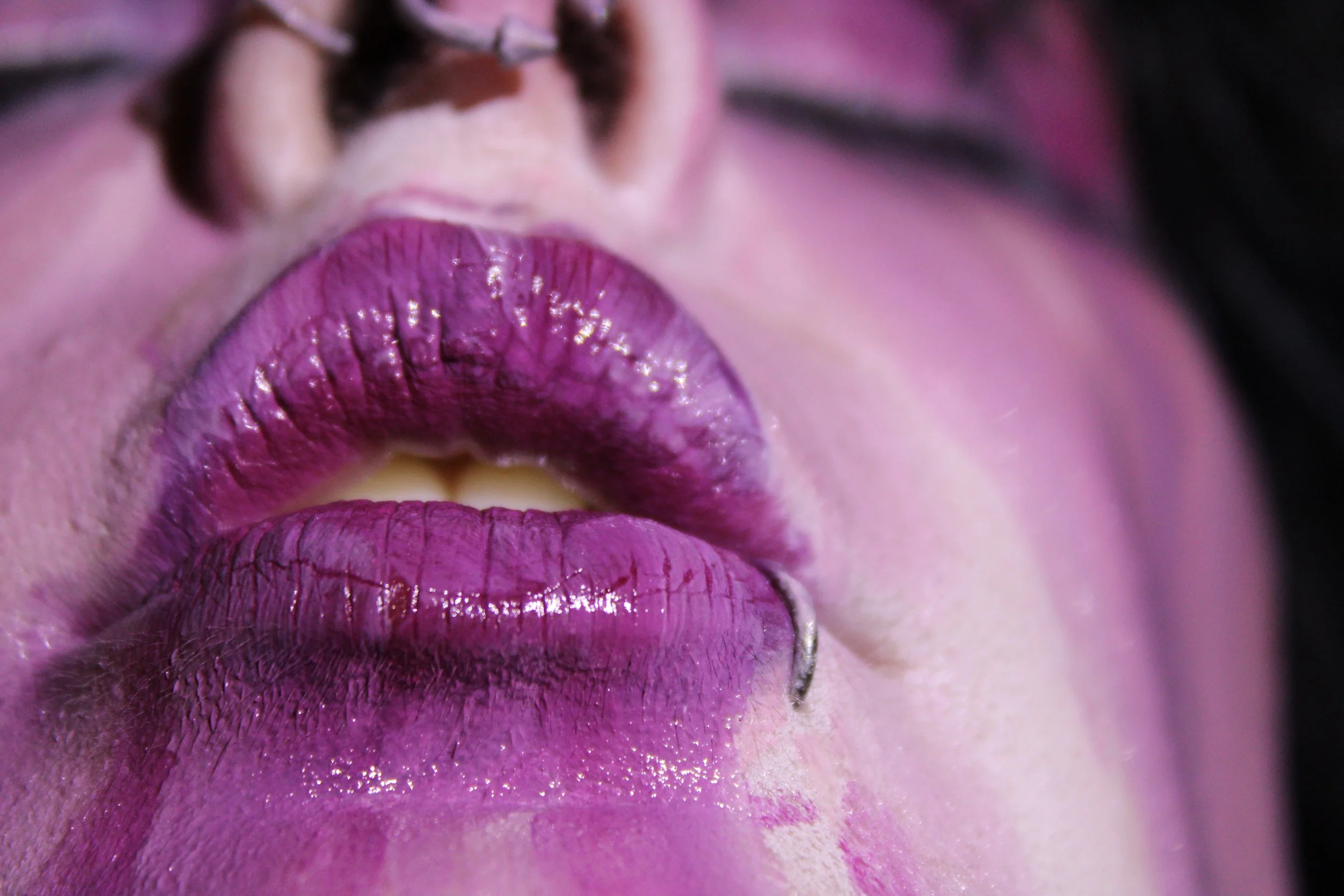 Close-up of a person's lips with purple lipstick, with part of their face visible, including their nose and some facial skin. The person's lips are glossy and prominent.