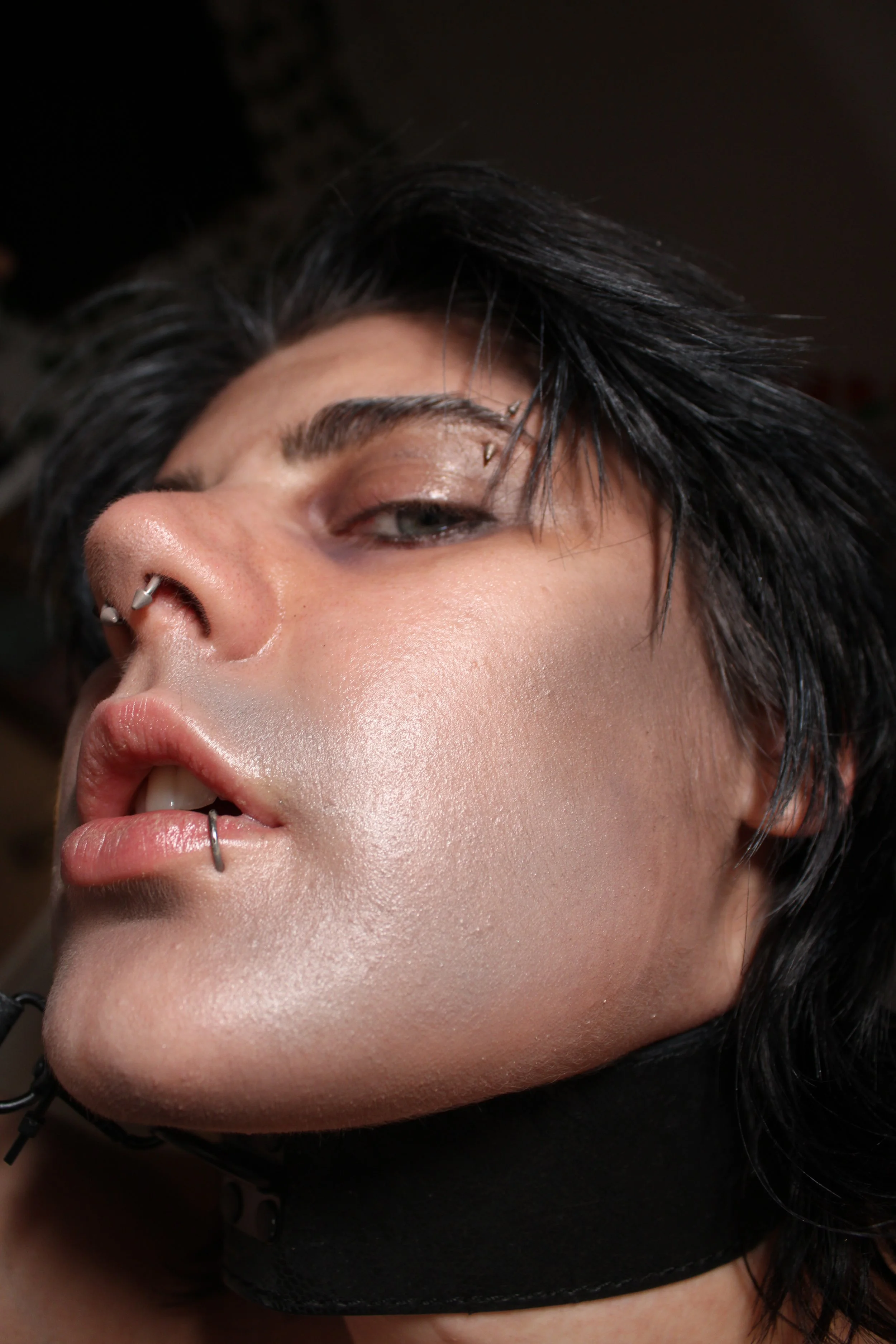 Close-up of a person with short black hair, multiple facial piercings, and makeup, wearing a black mesh face mask.