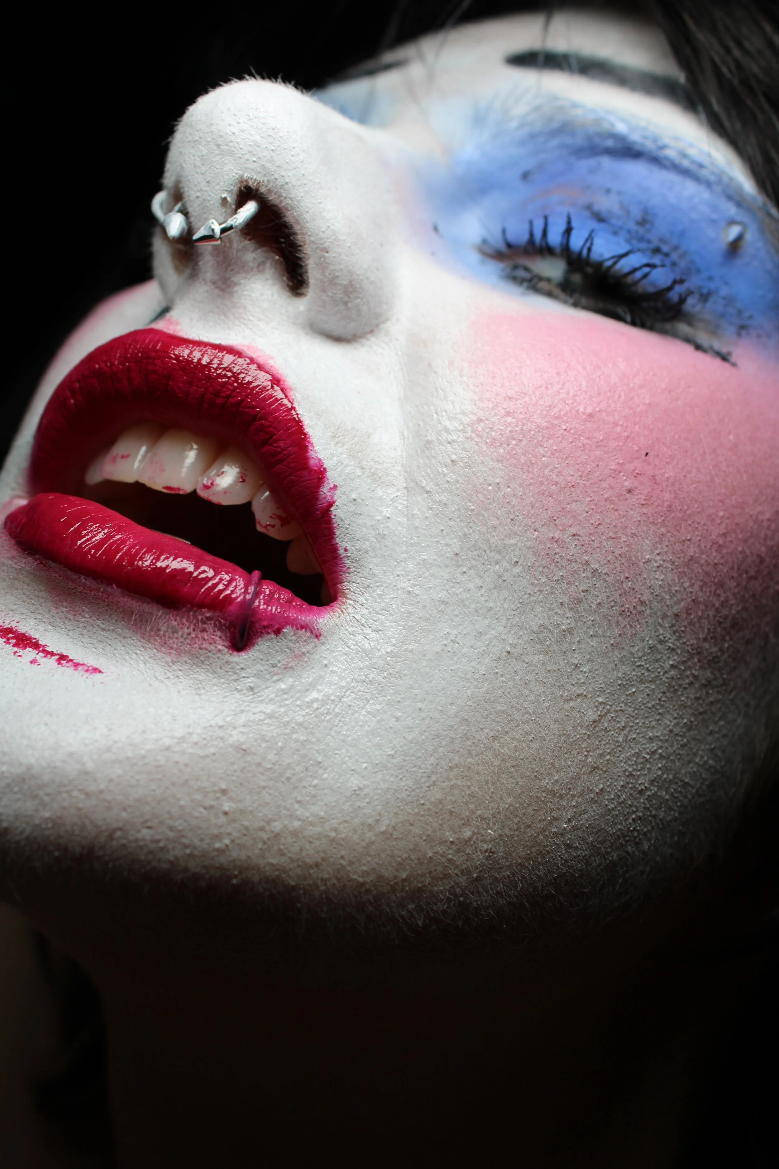 Close-up of a person with colorful makeup, including white face paint, bright red lipstick, and blue eyeshadow, with a nose piercing.