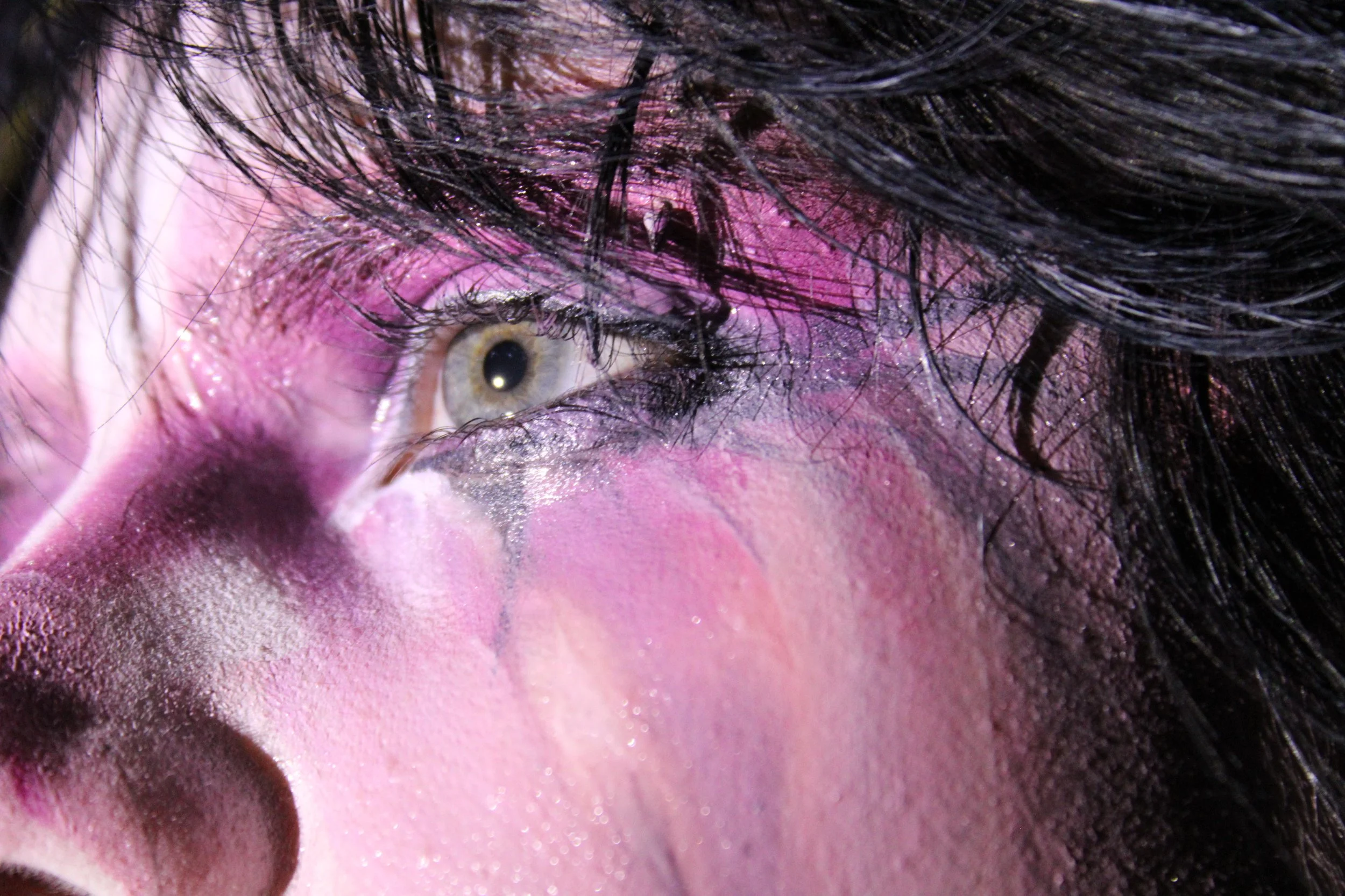 Close-up of a person's eye with colorful makeup, pink and purple eyeshadow, long dark eyelashes, and dark hair covering part of the face.