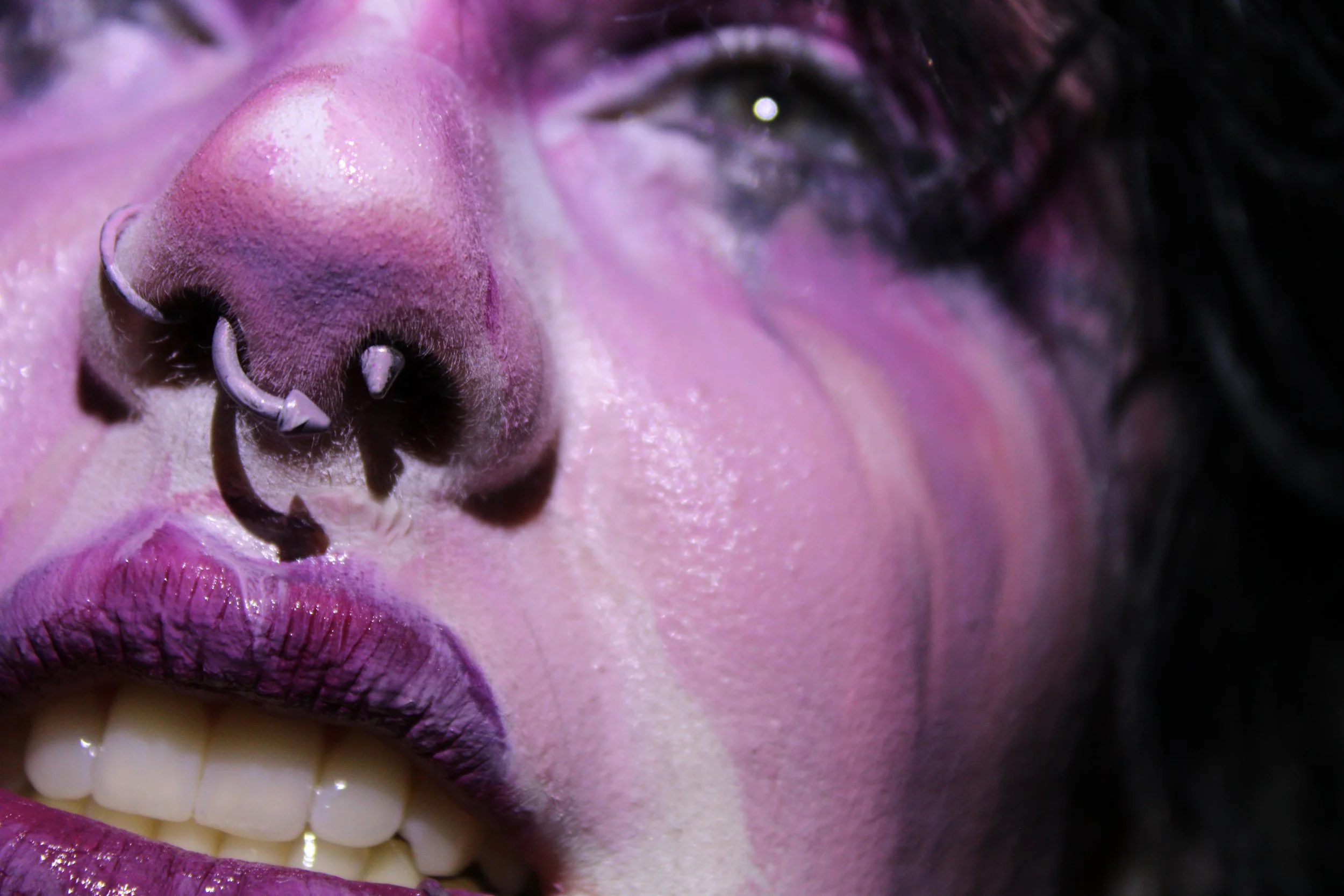 Close-up of a person's mouth with purple lipstick, showing teeth, lips, nose, and part of face, with piercings on the nose.