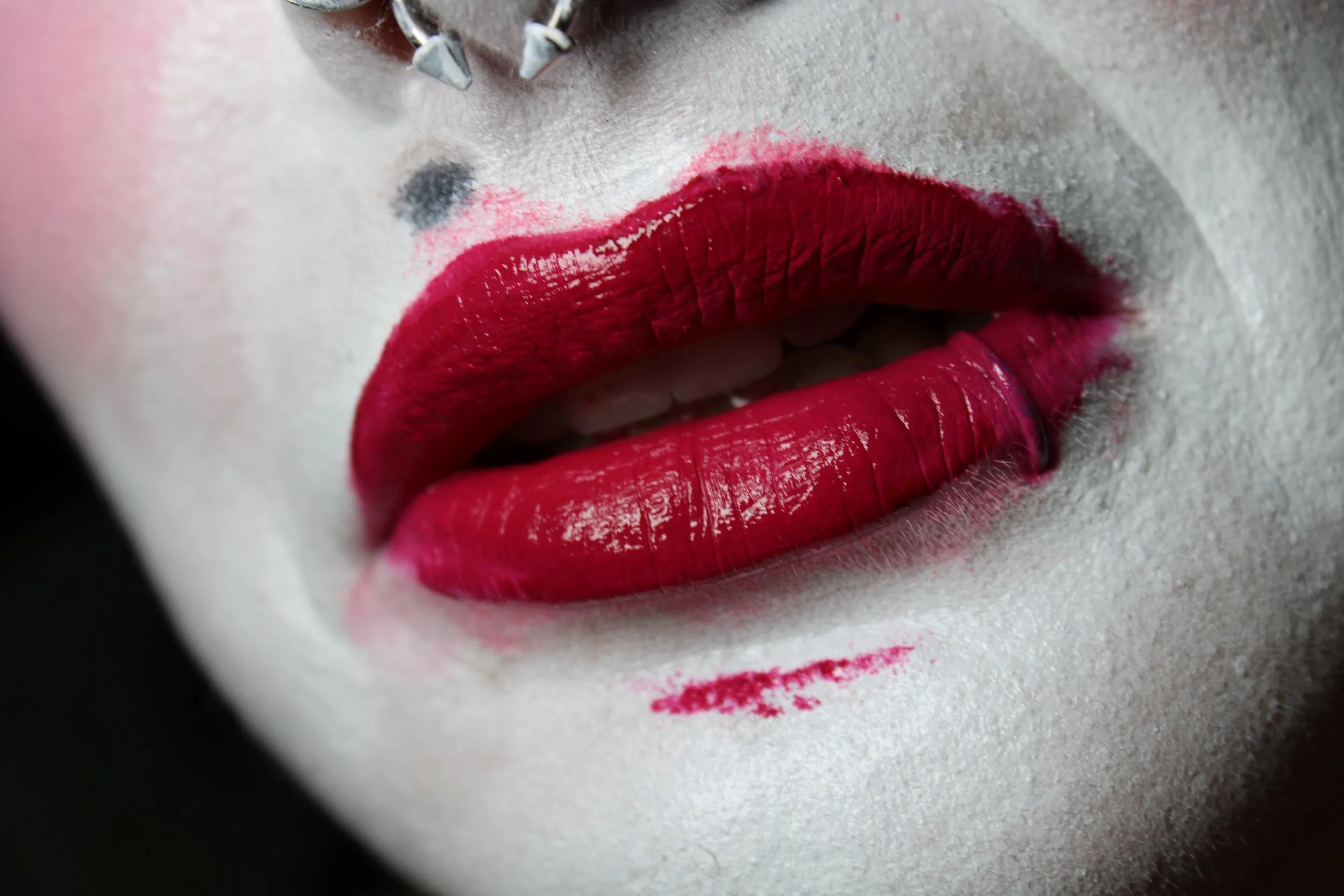 Close-up of a person's lips with bold red lipstick, white clown-like makeup, and there is a silver septum piercing.