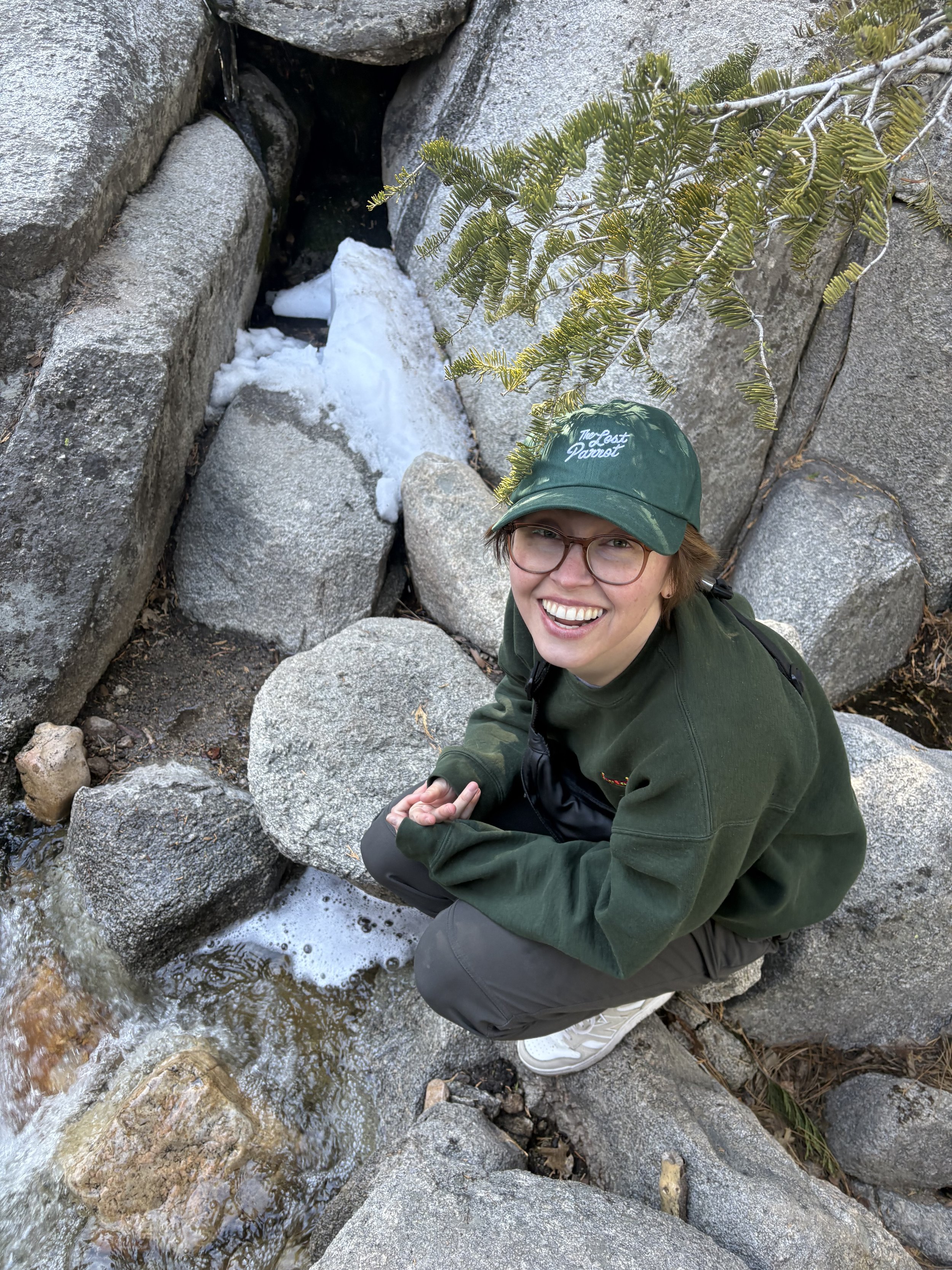 Person with green hat, green sweater, and grey sweatpants bending down by a waterfall and smiling.