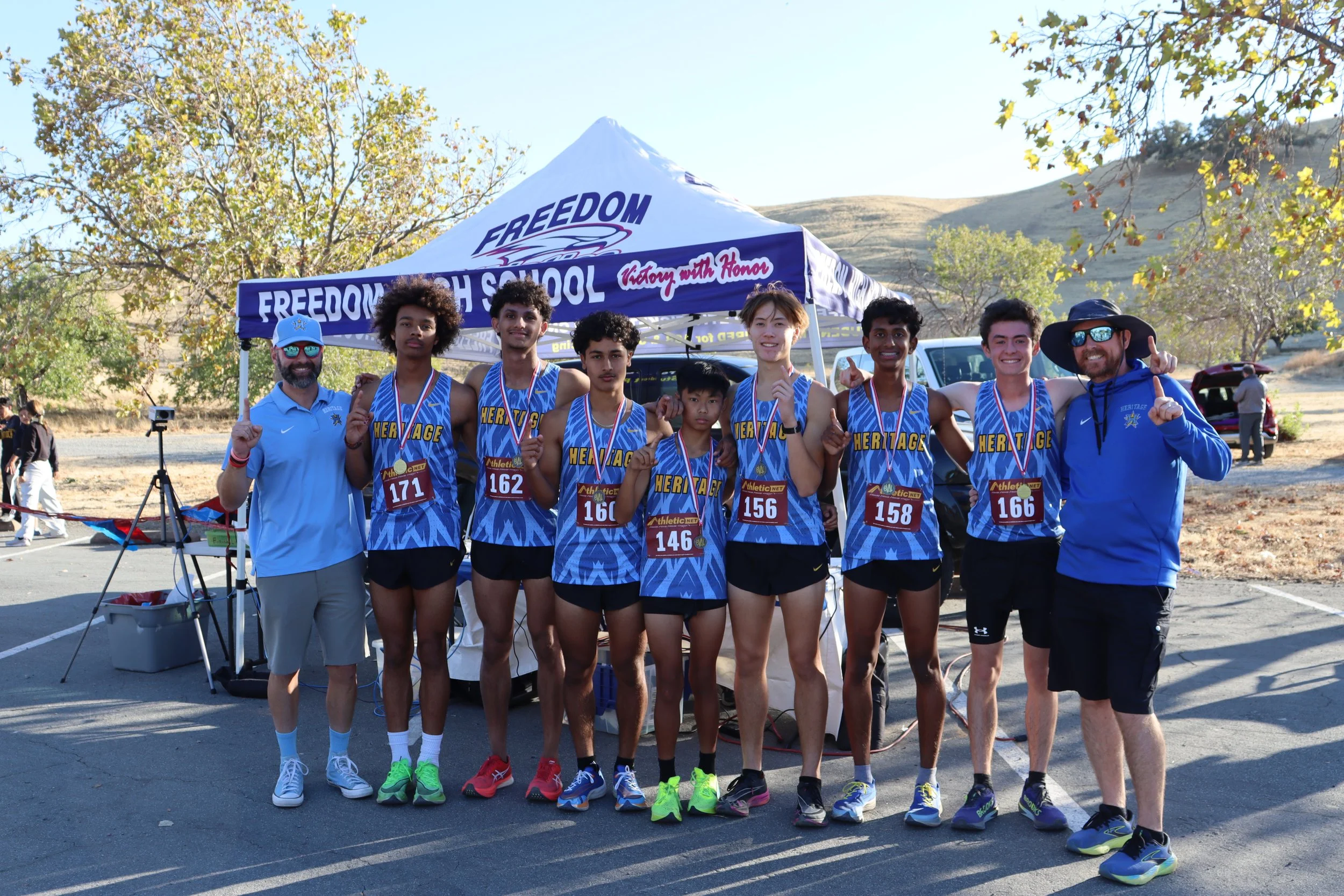 Group of young athletes from BEC and their coaches standing together after a race, under a blue canopy with 'FREEDOM HIGH SCHOOL' and 'Victory with Honor' printed on it, in an outdoor setting with trees and hills in the background.