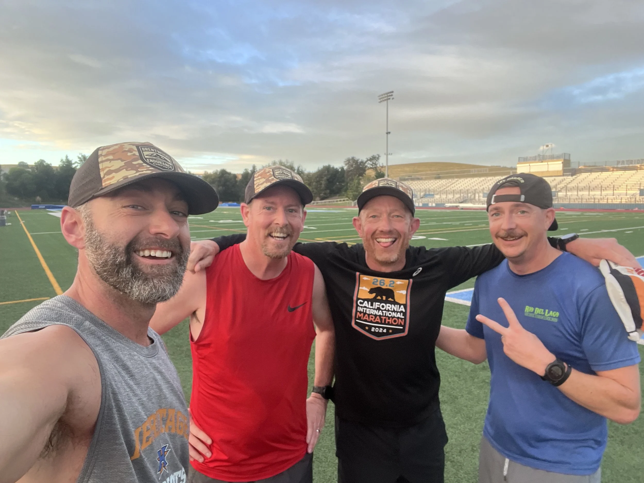 Brentwood Endurance Company Board members smiling and posing for a selfie on a sports field, with empty bleachers and a cloudy sky in the background, wearing athletic gear and caps.