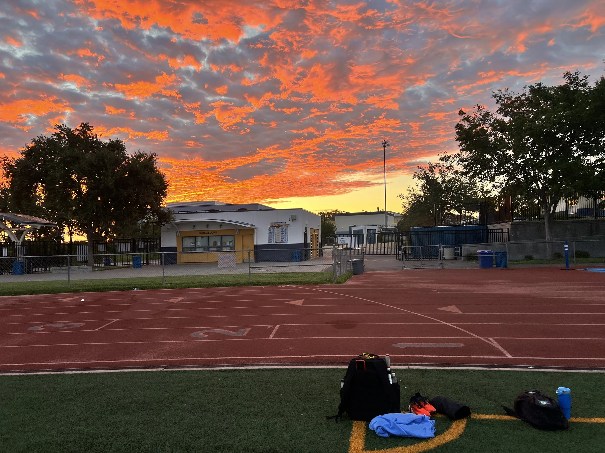 A view of Heritage High School's track field at sunset with orange and gray clouds in the sky. Several trees and a small building are visible in the background. Items are scattered on the field, including clothing and bags.
