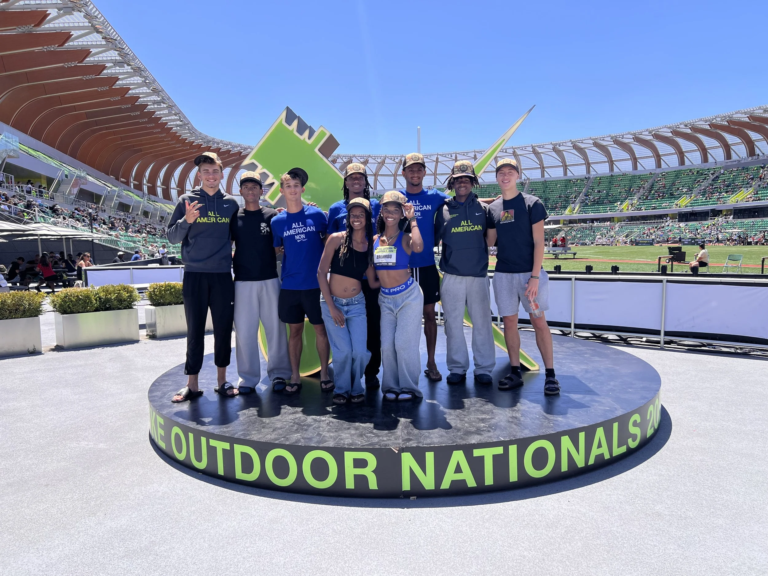 Group of diverse young athletes standing on a circular stage at the Outside Lands music festival, Golden Gate Park, San Francisco, with a large green and black stage decoration behind them and stadium seating filled with spectators in the background.