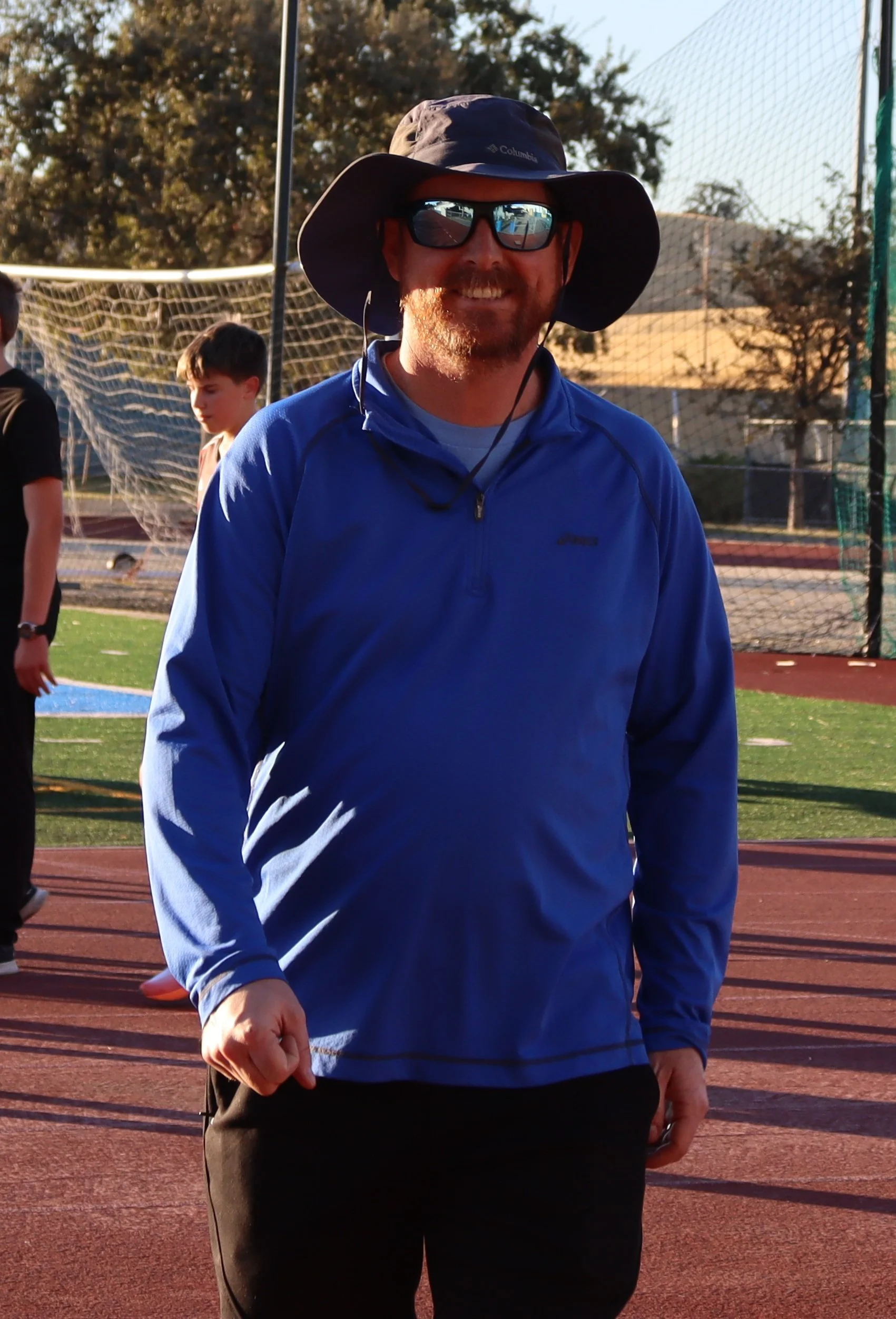 Smiling man in blue hoodie with backpack standing on a running track at an outdoor sports field.
