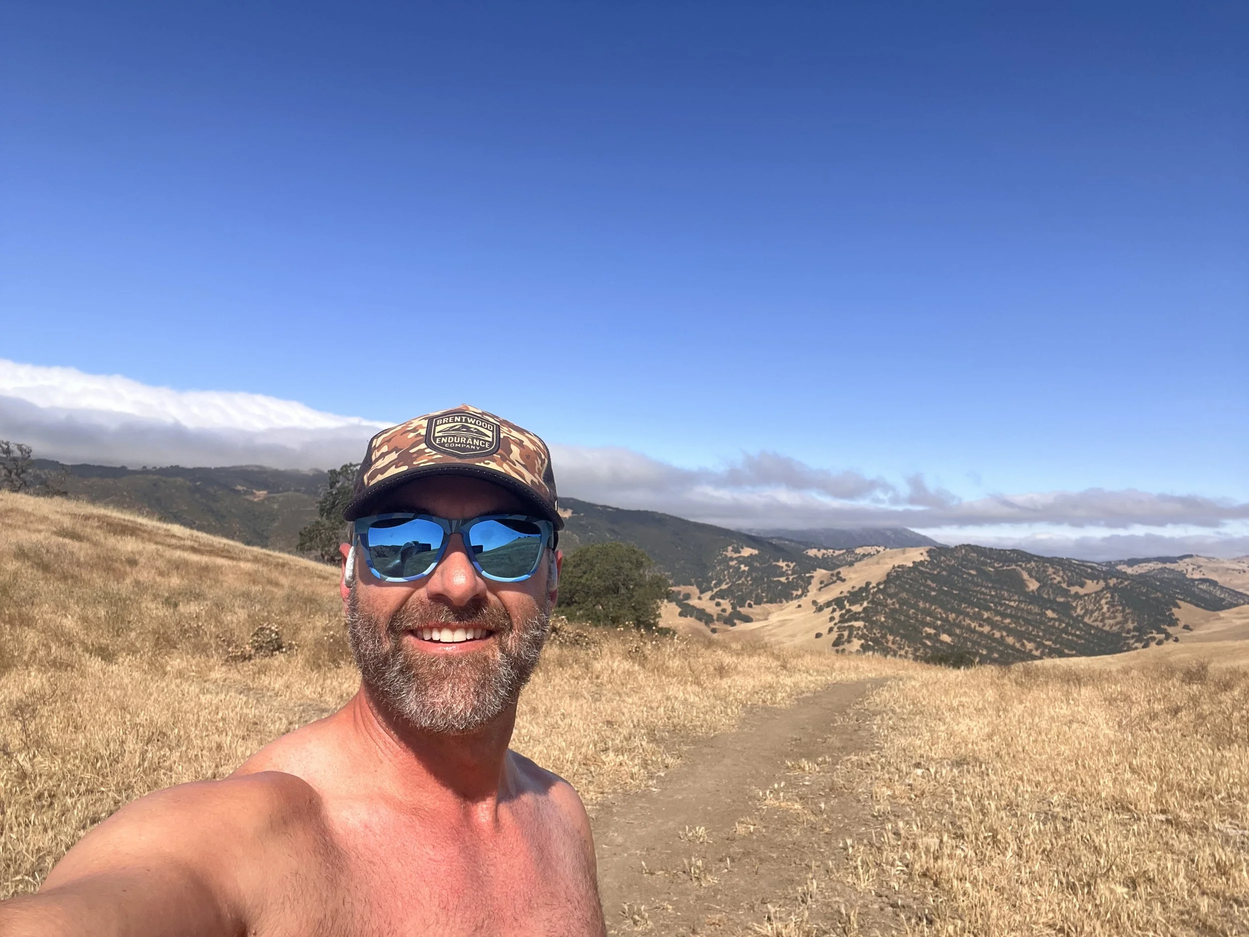 A man taking a selfie outdoors in a hilly, grassy landscape under a clear blue sky. He is wearing sunglasses, a camo baseball cap with a logo, and is shirtless, smiling at the camera.