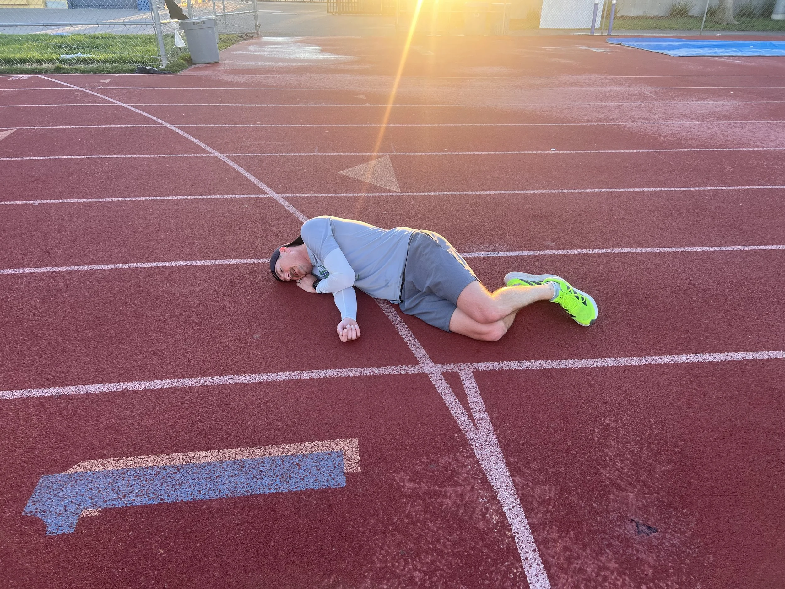 A man lies on the running track exhausted after a great performance, wearing a gray long sleeve shirt, gray shorts, bright yellow shoes, and a black headband, with the sun setting in the background.