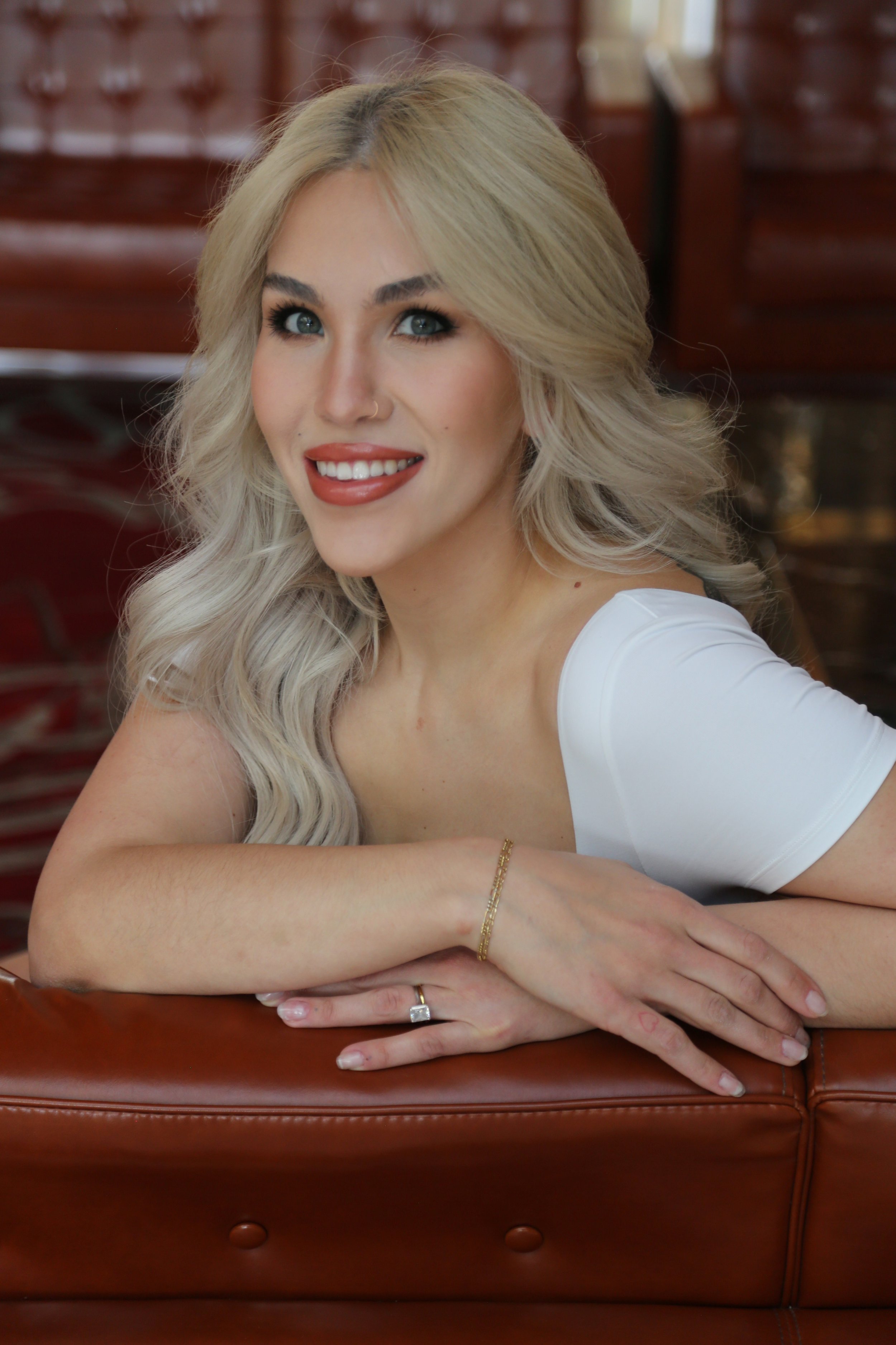 A woman with long, wavy blonde hair, light skin, and blue eyes, smiling with red lipstick, resting her arms on a brown leather couch.