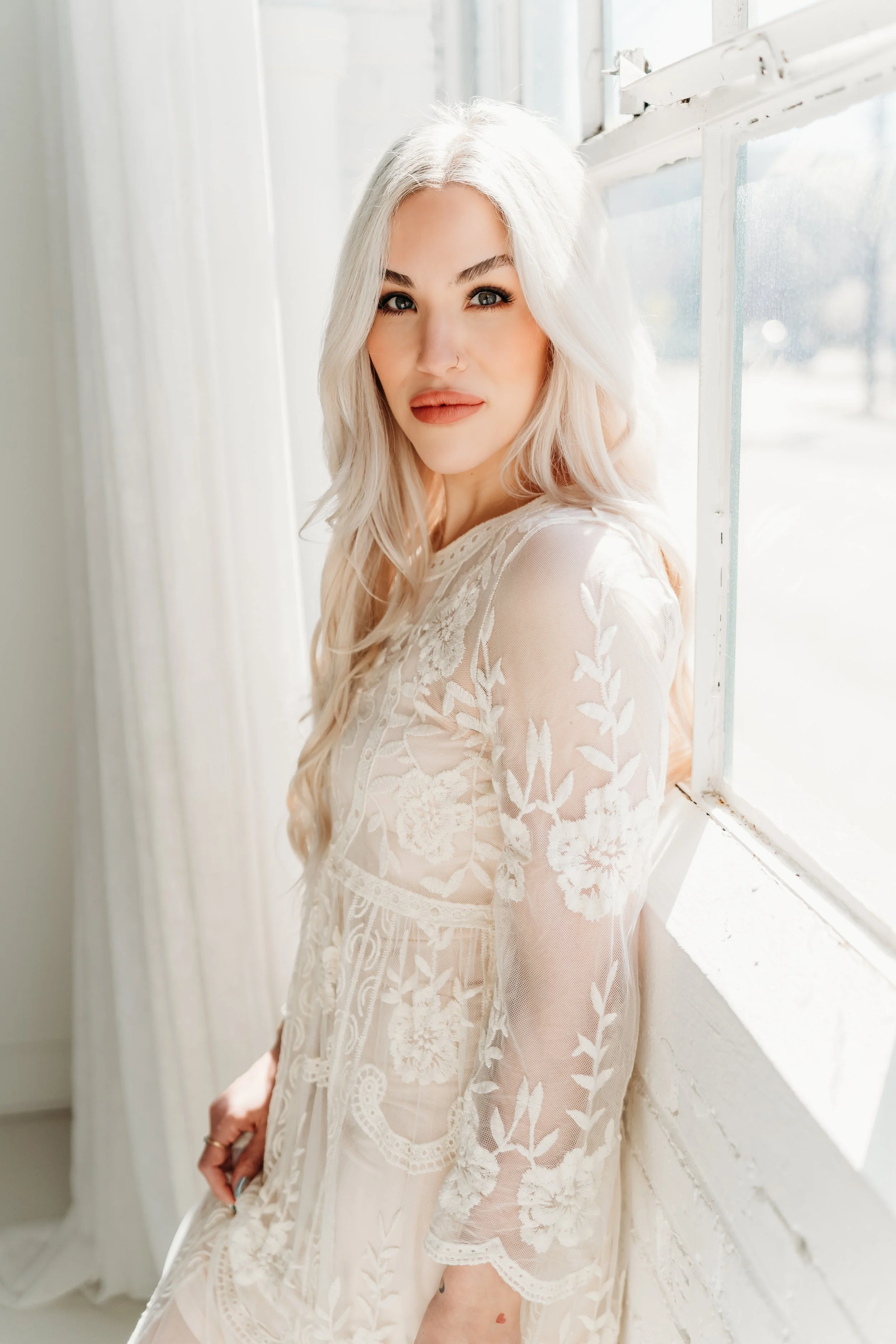 A woman with long blonde hair and light skin standing by a window, wearing a sheer lace dress with floral patterns, looking at the camera with a neutral expression.