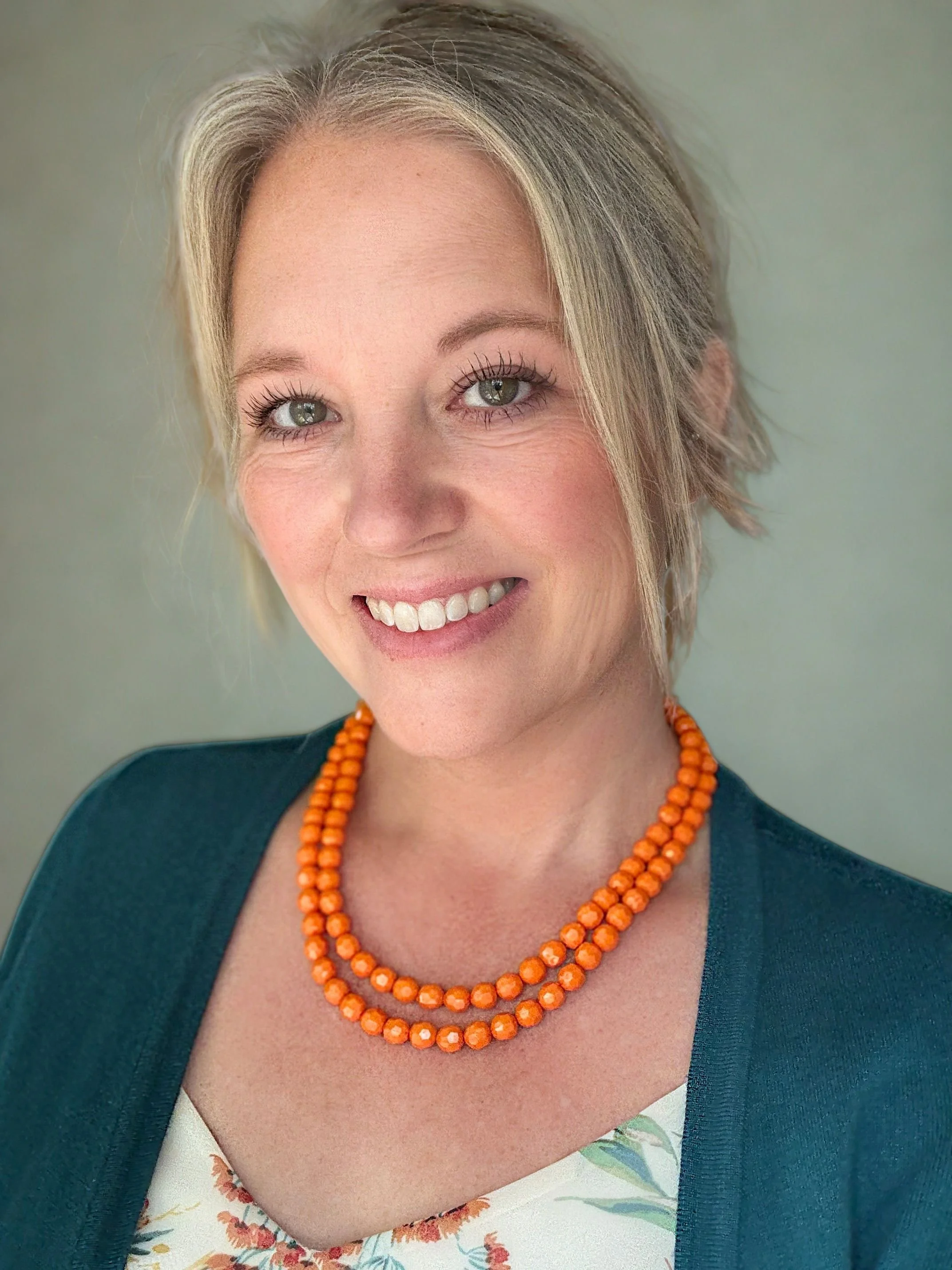 A woman with short blonde hair, green eyes, and a bright smile wearing a dark teal blazer over a floral top, accessorized with two orange beaded necklaces.
