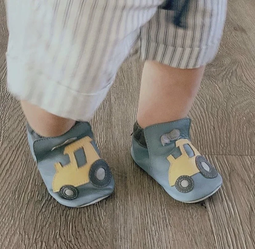 Close-up of a child's legs and feet wearing blue shoes with yellow construction vehicles on the toes, standing on a wooden floor. I designed the motifs on the shoes.