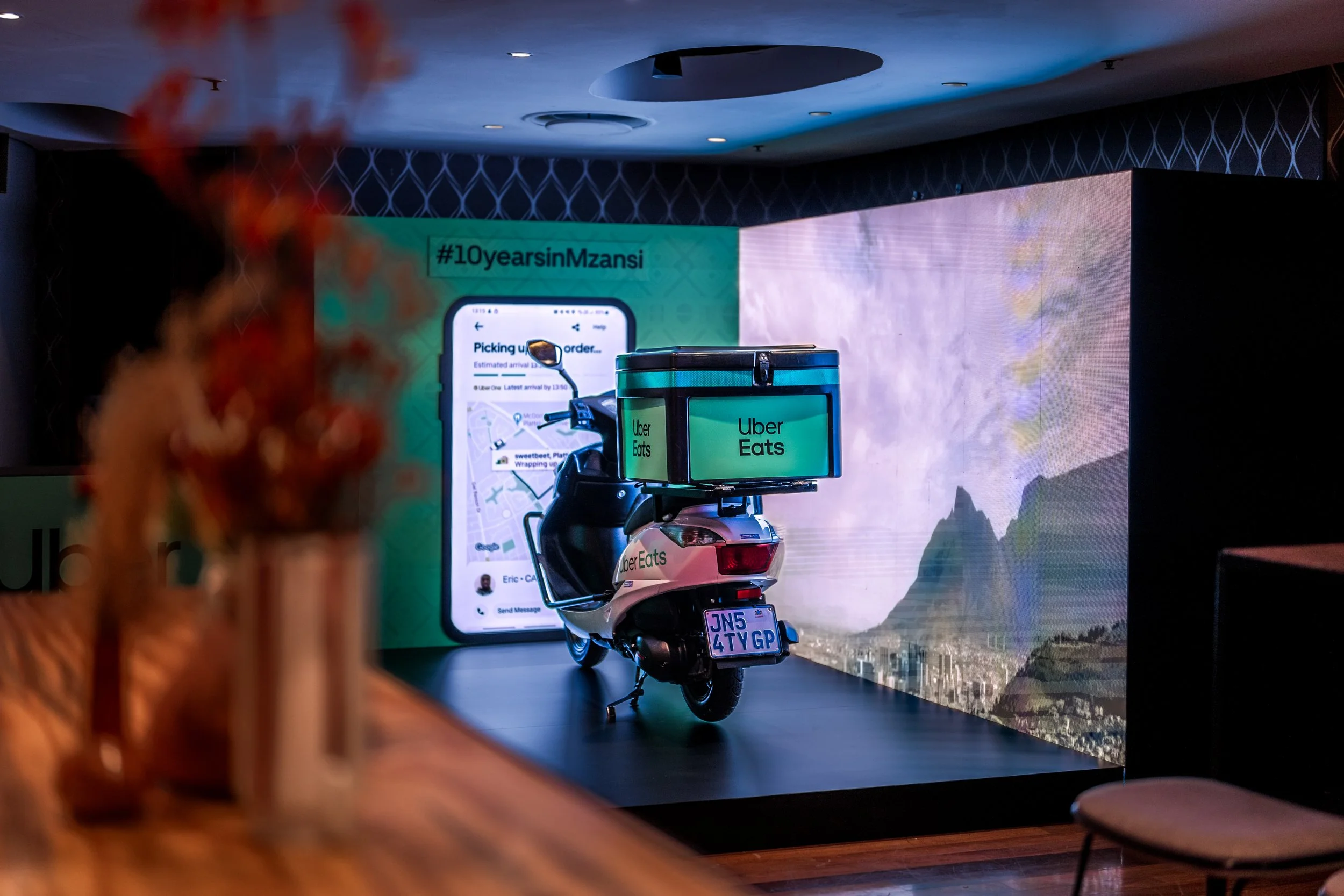 A display with a large digital screen, a scooter with Uber Eats branding, and a poster for Uber Eats, with hashtags and a map showing delivery details.