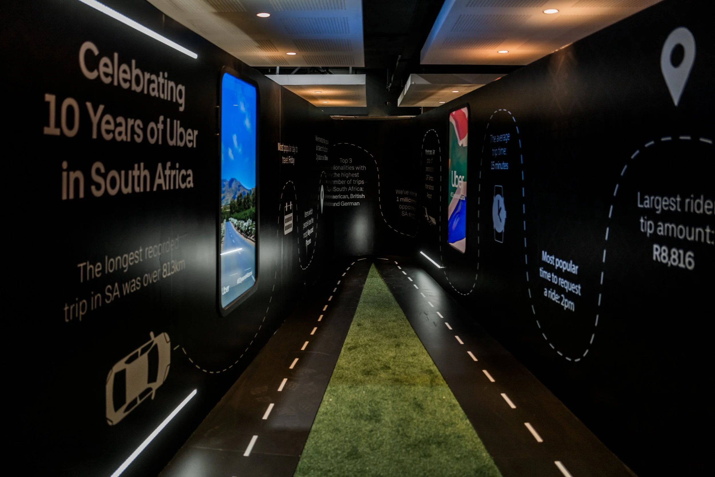 A dark hallway exhibit celebrating 10 years of Uber in South Africa with informational displays, digital screens, and illuminated road markings on the floor.