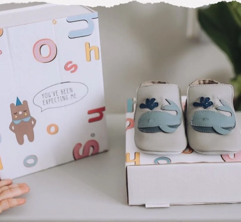 A pair of baby shoes with whale designs sit on top of a white shoebox, next to a larger box with cartoon bear and alphabet decorations. I designed the motifs.