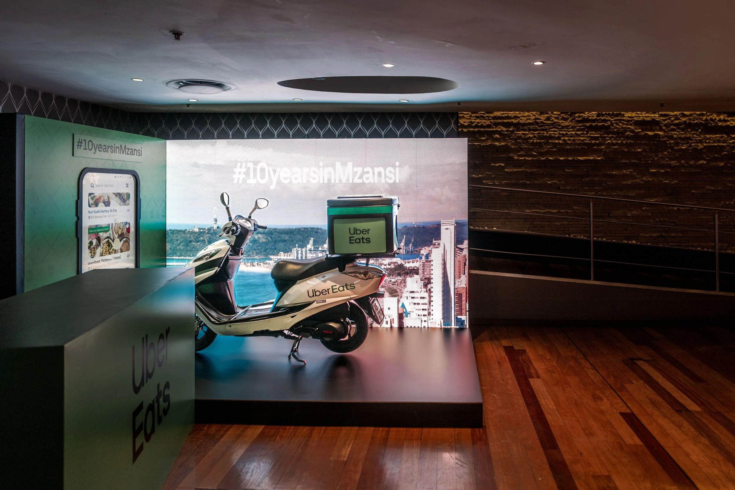 A scooter with an Uber Eats delivery box parked indoors near a large cityscape digital display. The display has the hashtag #10yearsInMzansi and branding related to Uber Eats.