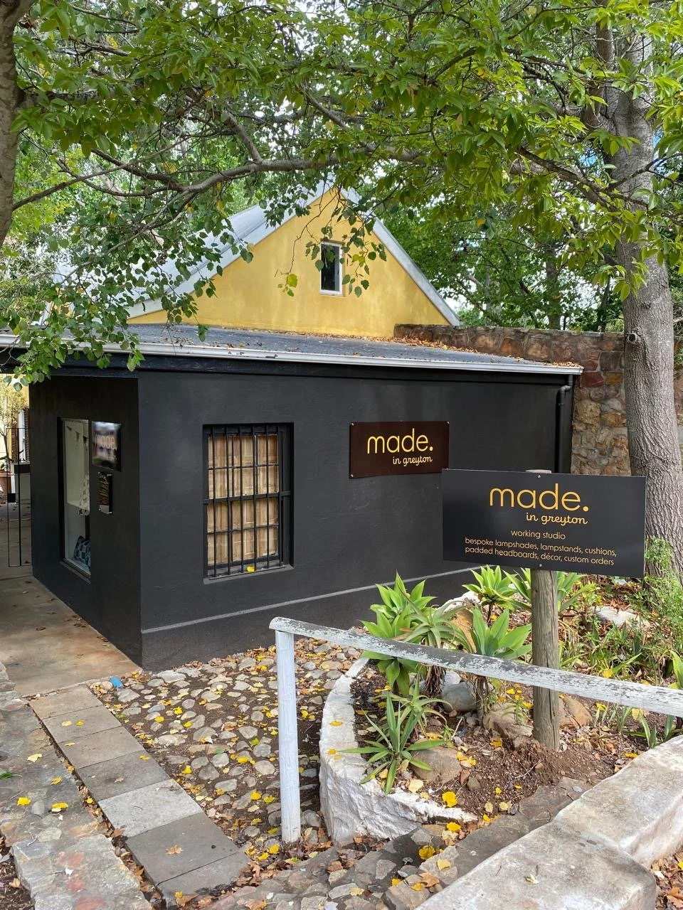 A small black building with signs reading "made. in greyton" and information about being a working studio for lampshades, lampstands, cushions, and custom orders, situated in a garden with plants and a winding stone path, surrounded by trees and a ye