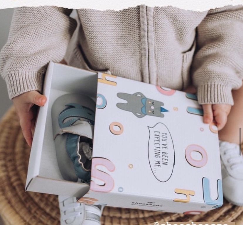 Child in beige sweater holding a gift box with a pair of baby shoes inside, and a lid with a cartoon bunny and colorful shapes. I designed the motifs.