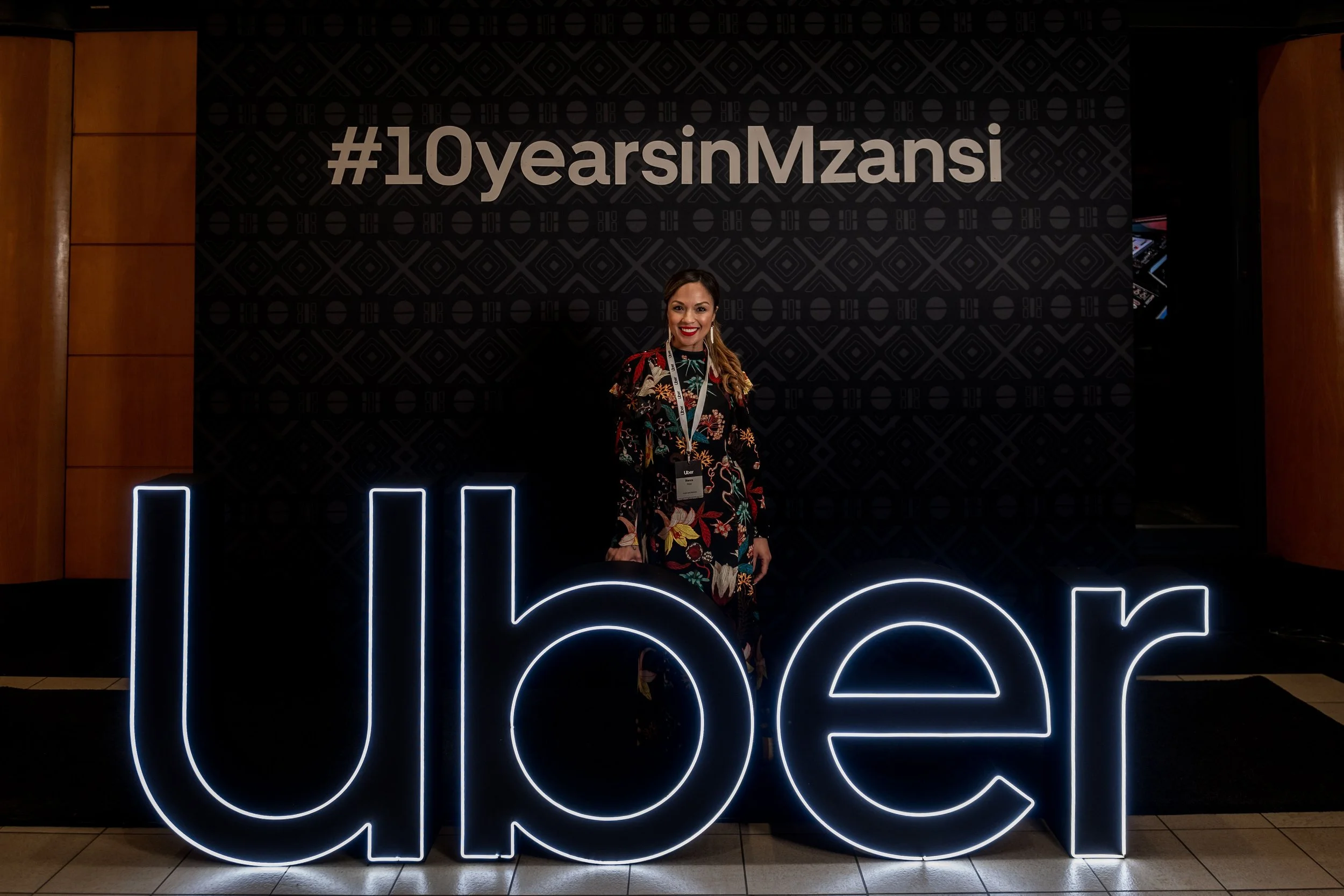 Woman standing in front of a wall with illuminated large text celebrating 10 years for Mzansi, with a hashtag #10yearsInMzansi above her.