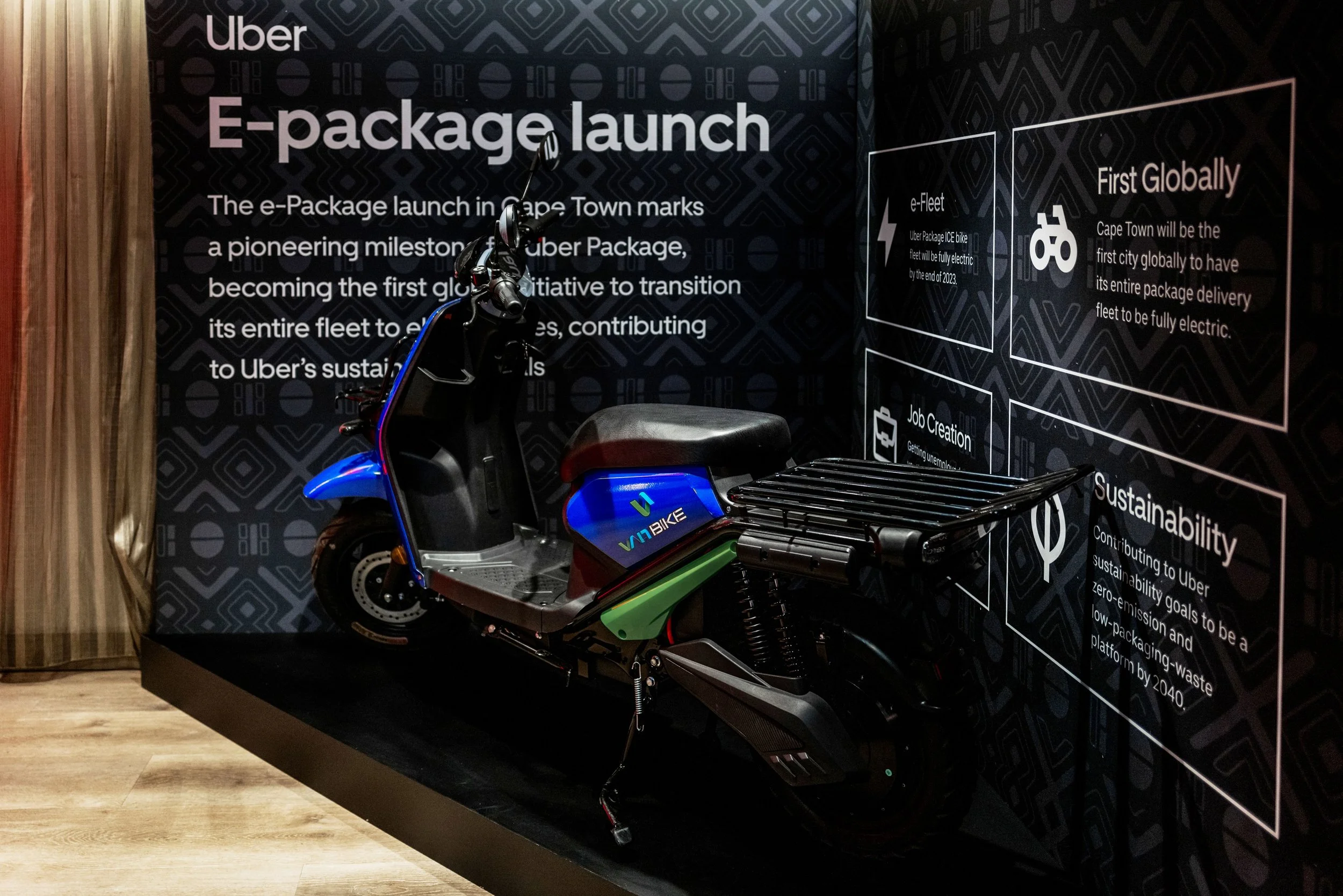 An electric scooter displayed on a black platform at a showcase event for Uber's e-package launch in Cape Town, with informational banners in the background.