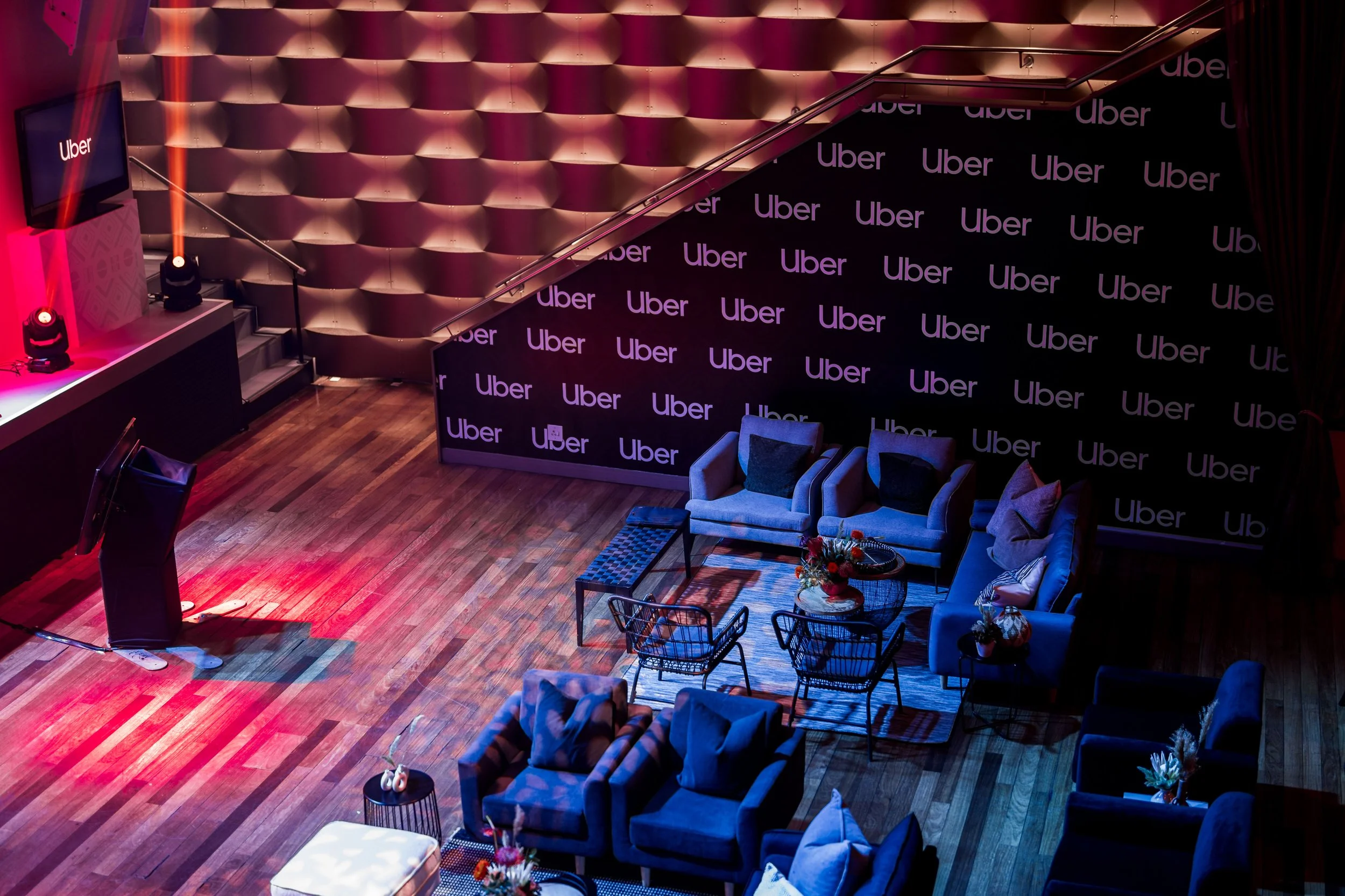 An indoor event space with couches, chairs, and a small table arranged on a wood floor. There are red and blue lighting, a large black wall with the Uber logo repeated, a stage with spotlights, and a television displaying the Uber logo.