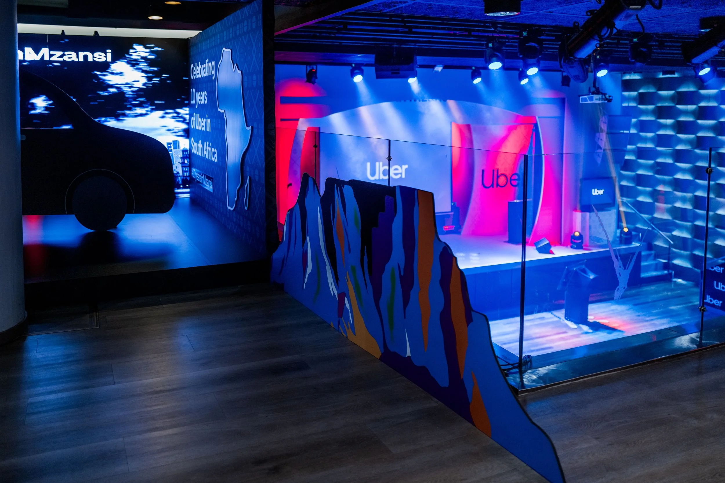 A dark indoor event space with vibrant blue and pink lighting. A large wall displays a digital graphic with a silhouette of a person's face, text mentioning Uber, and a map outline of South Africa. There are stage lights and equipment hanging from the ceiling, and a small barrier with colorful artwork in the foreground.