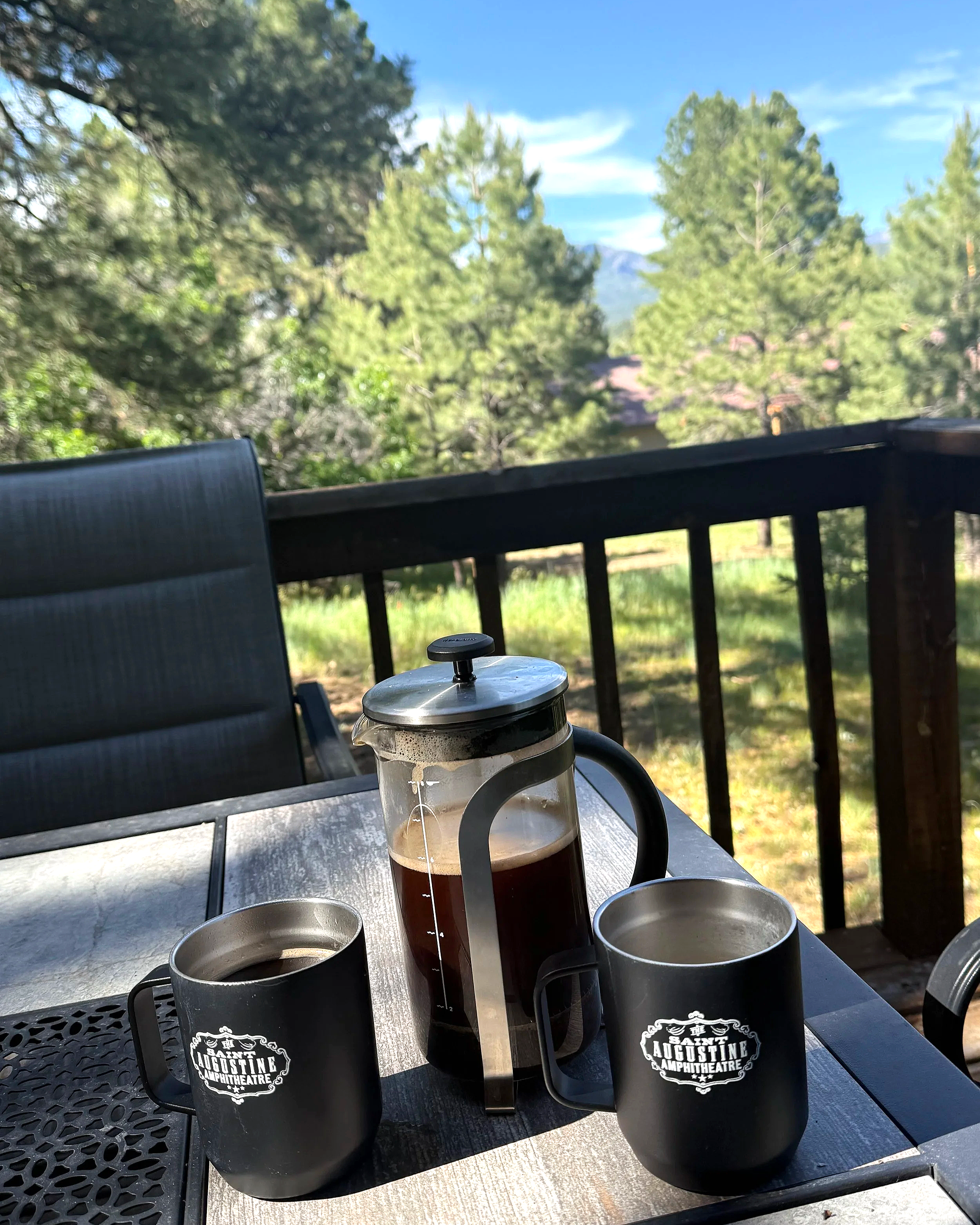 Outdoor patio table set with a French press filled with coffee and three black mugs with a logo reading 'Saint Augustine Amphitheatre,' surrounded by trees and a railing in the background.