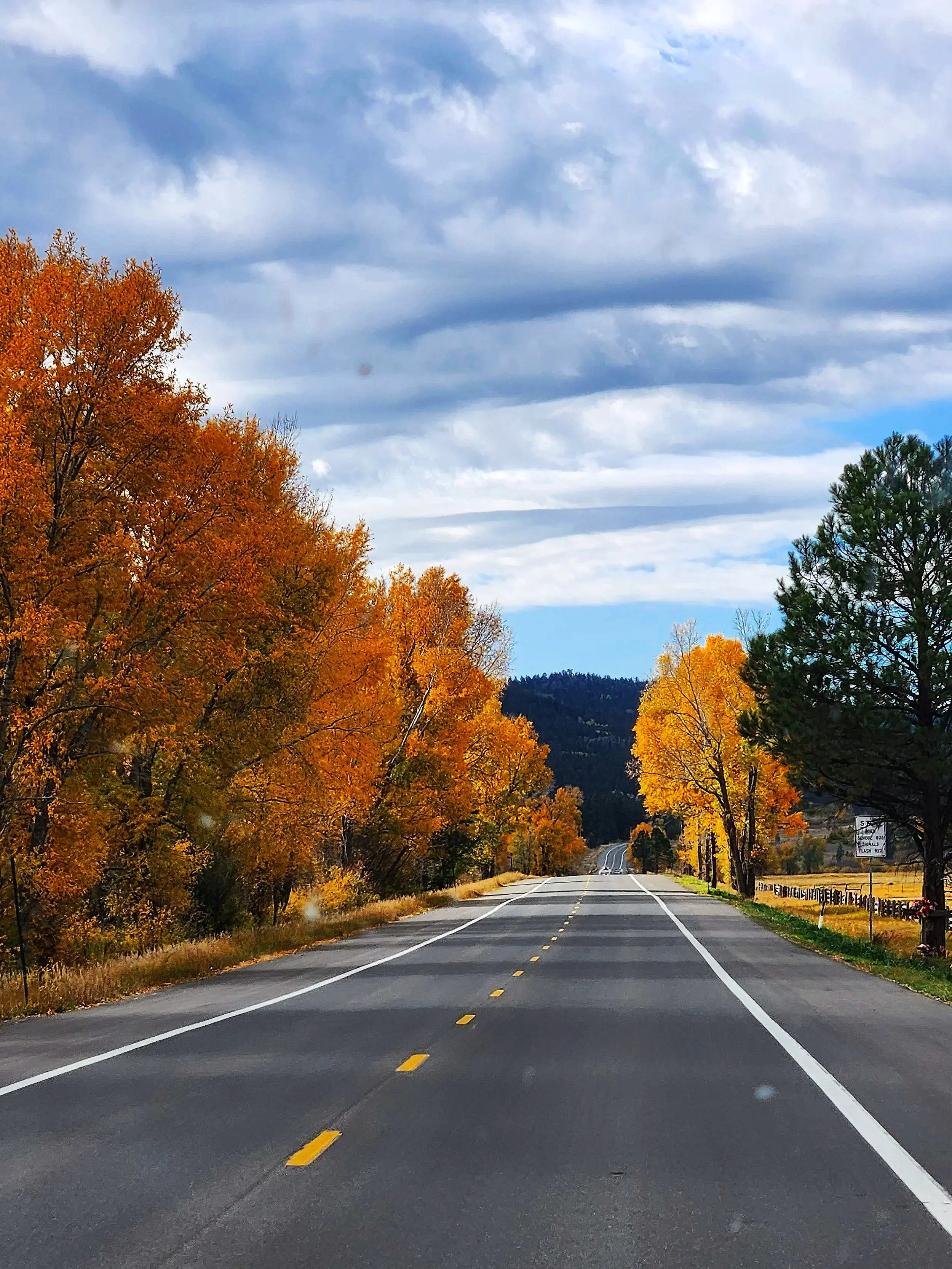 A two-lane road with a dashed yellow center line and solid white edge lines, surrounded by colorful autumn trees with orange, yellow, and green leaves, under a partly cloudy sky.