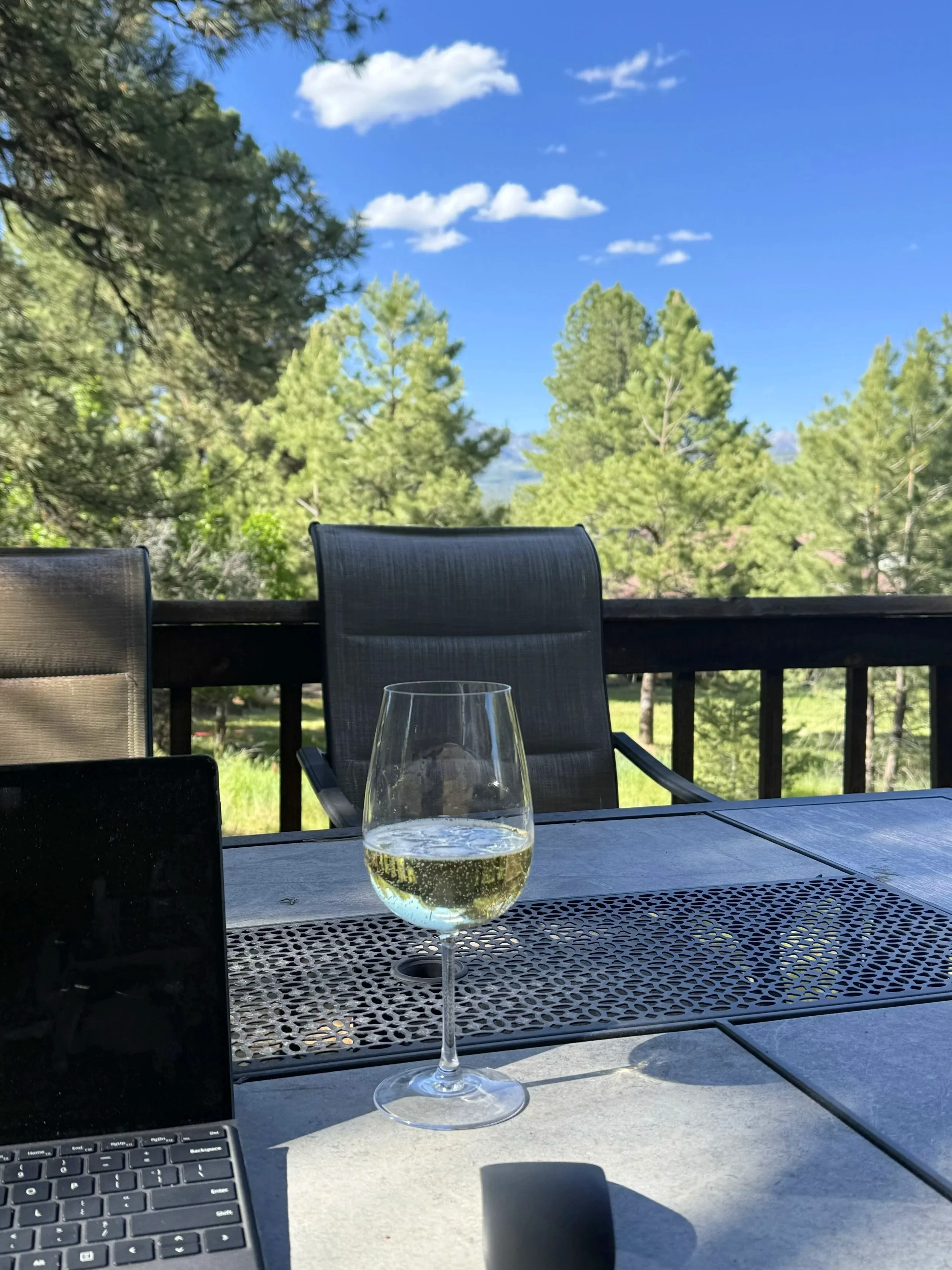A glass of white wine on an outdoor table with a tablet and laptop, overlooking a landscape with green trees and a blue sky with white clouds.