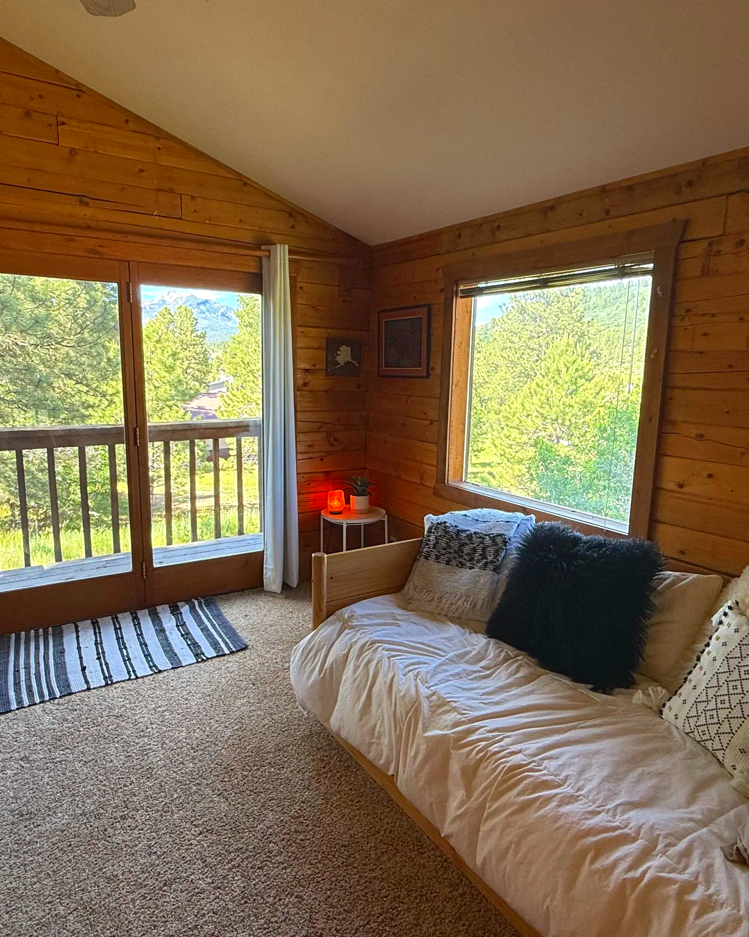 A bedroom with wooden walls and large windows showing green trees and mountains outside. There is a bed with a white blanket and black and white pillows, a small white side table with a lamp and a plant, and sliding glass door with a curtain. A strip