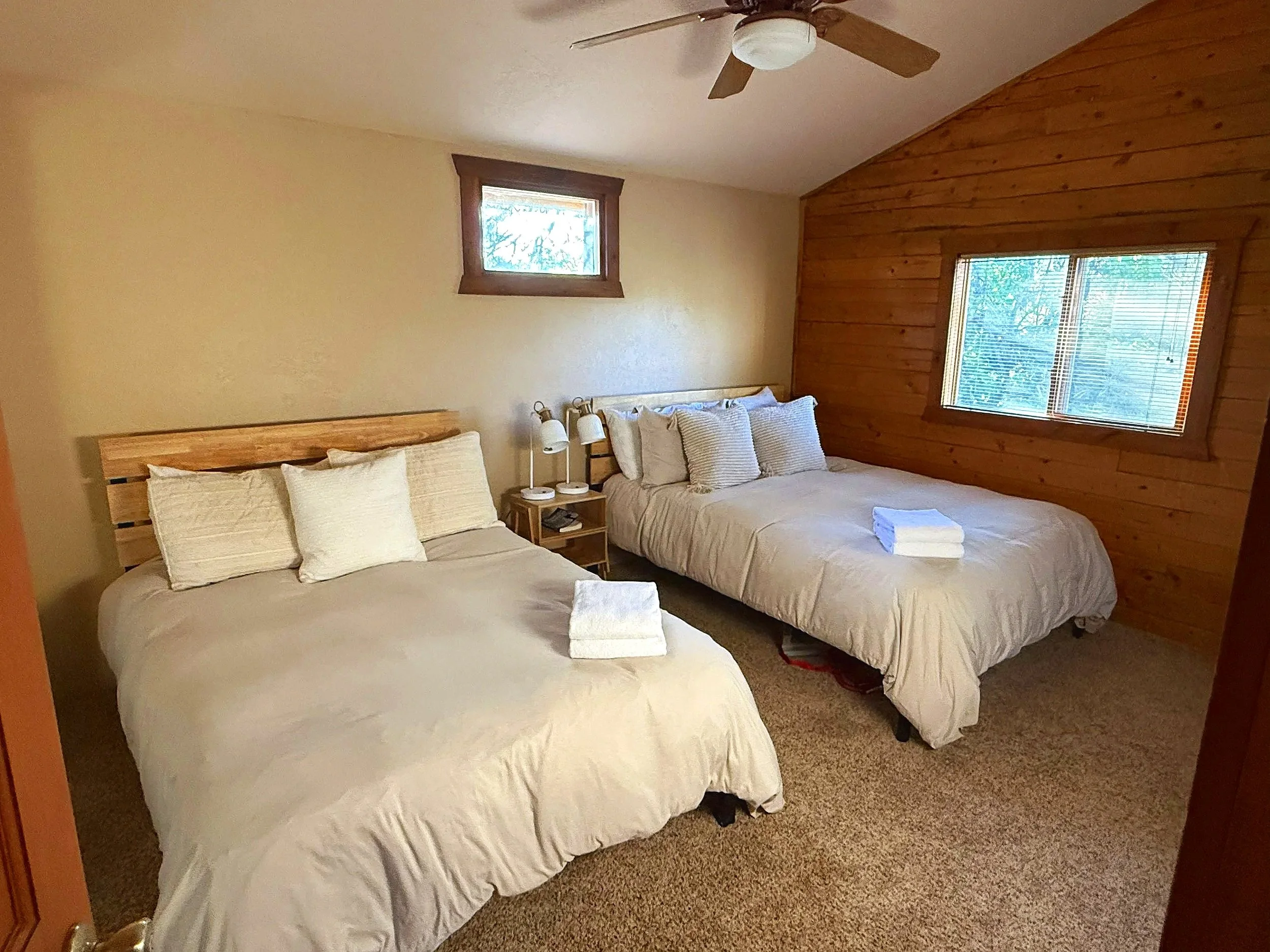 Two beds with white bedding and multiple pillows, a small wooden bedside table with two white lamps and a phone, two towels on each bed, a window with blinds, and a wooden wall with a small window.