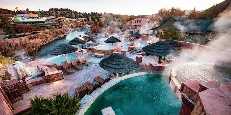 Outdoor hot springs with steaming water, wooden lounge chairs, and thatched umbrellas, set along a river with buildings and trees in the background.