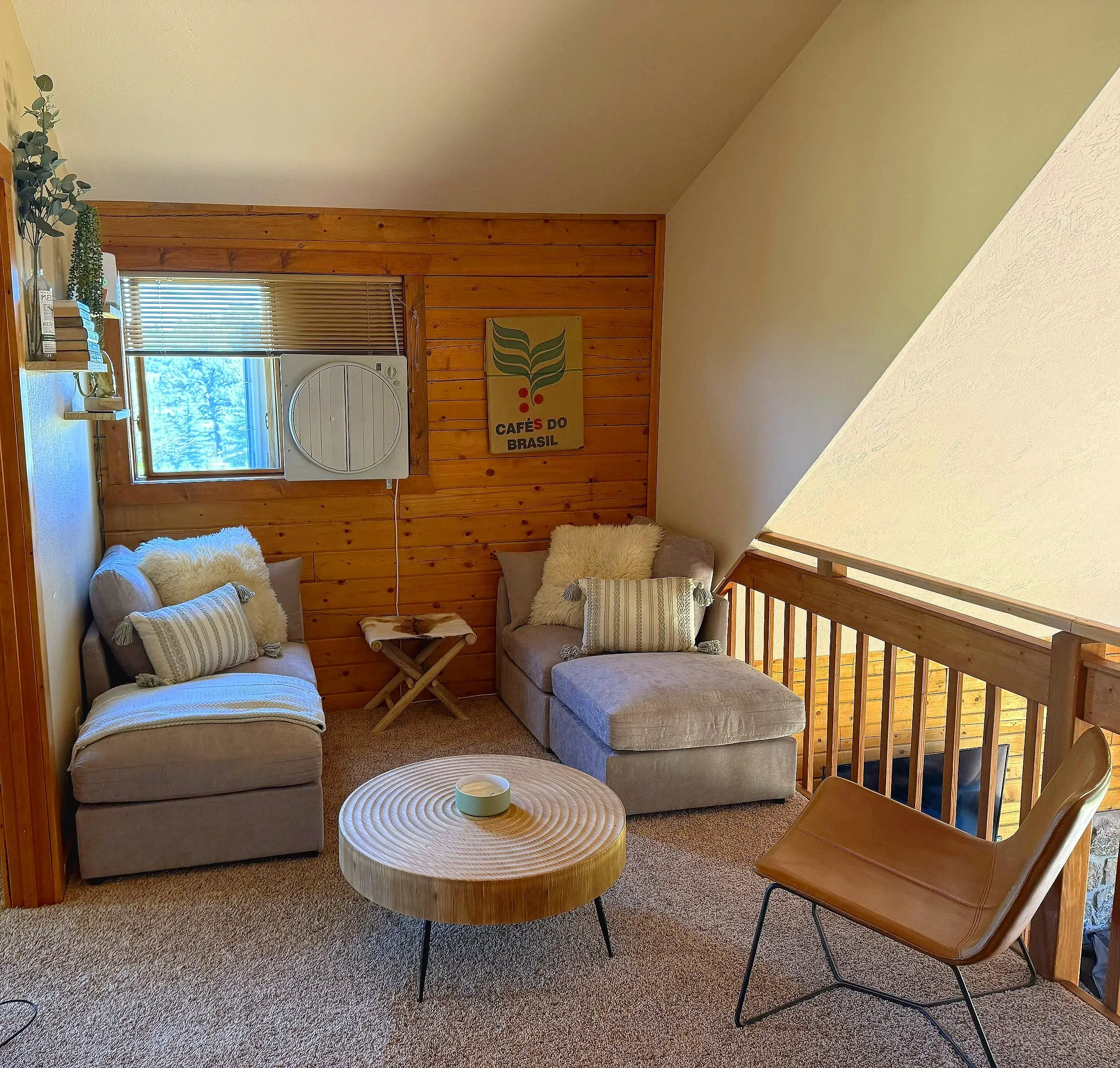 Living room with two sofas, round wooden coffee table, wooden chair, window with blinds, wood-paneled wall, laundry dryer, plant, and framed art that says "Café do Brasil."