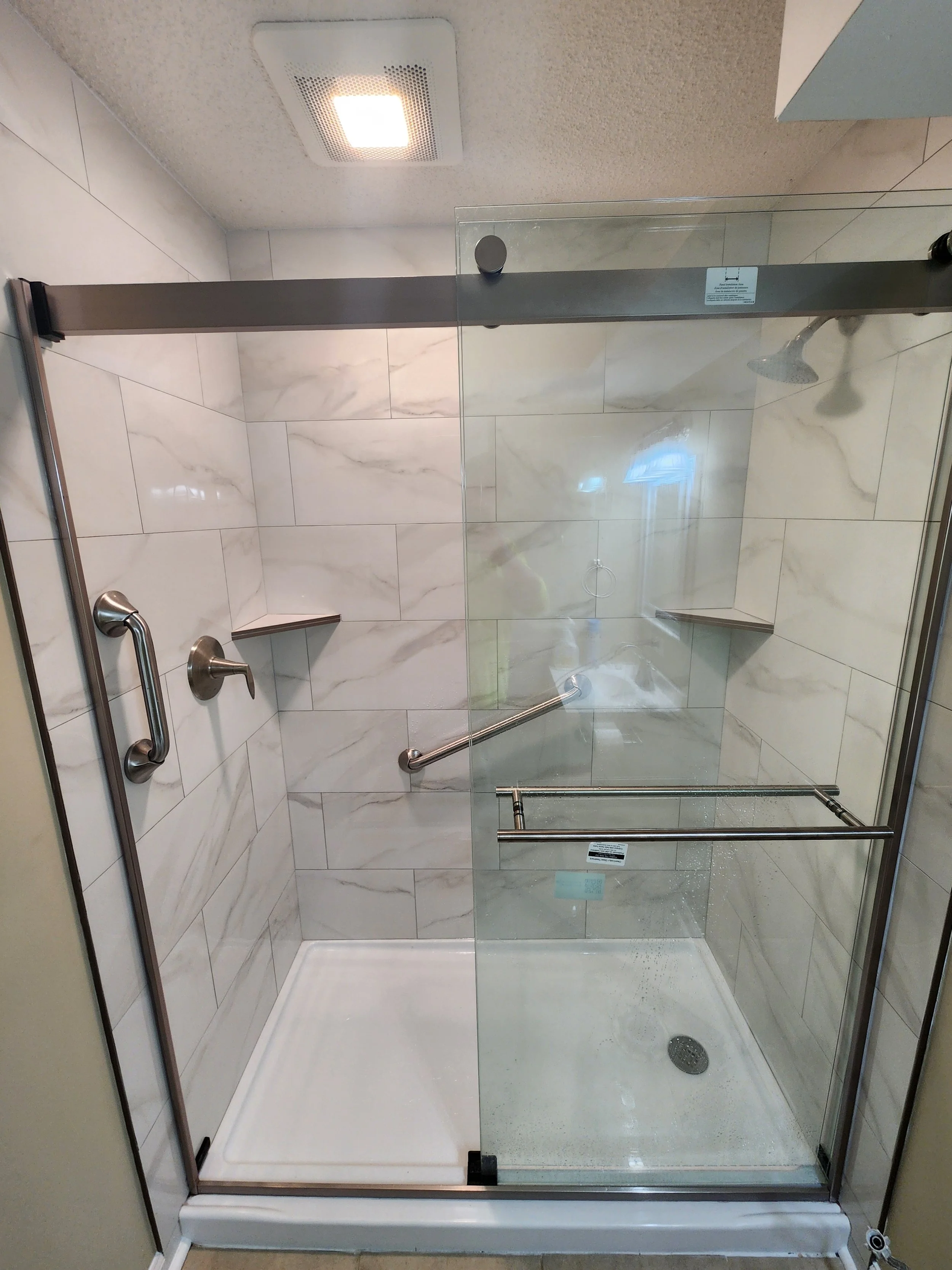 A modern walk-in shower with a glass door and marble tiles, featuring a handheld showerhead, a wall-mounted showerhead, grab bars, and a white shower base.