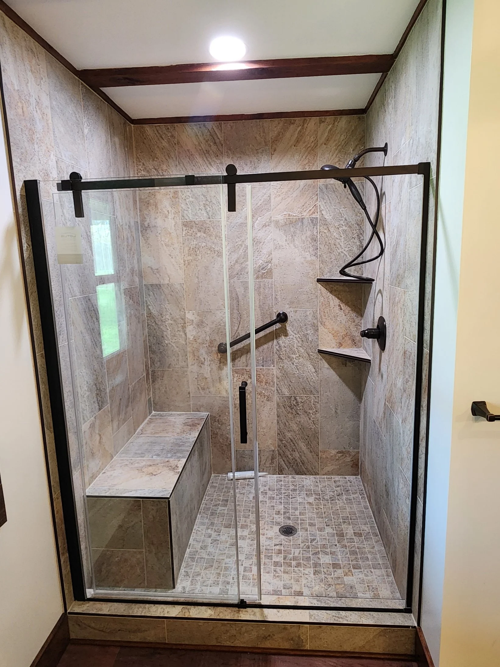 A tiled shower with a glass sliding door, built-in bench, a wall-mounted shower head, a small corner shelf, a window, and a drain on the shower floor.