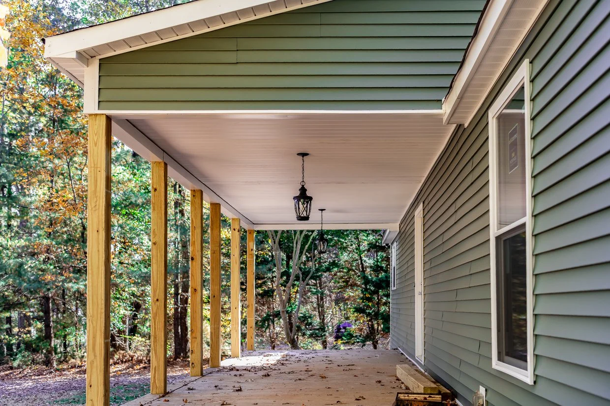 Under construction porch with new wooden support posts and two hanging lanterns, attached to a house with green vinyl siding, overlooking a wooded area.